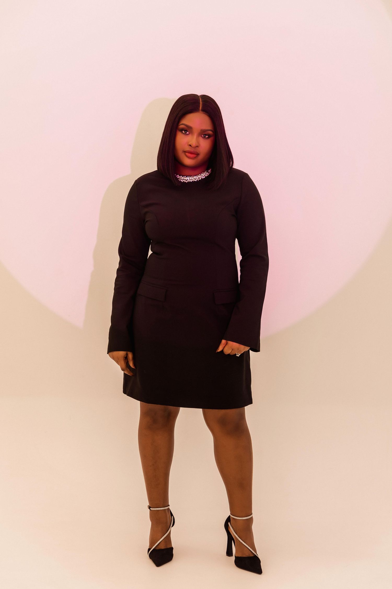 Woman in black long-sleeve mini dress with rhinestone collar and strappy heels poses against pink background.