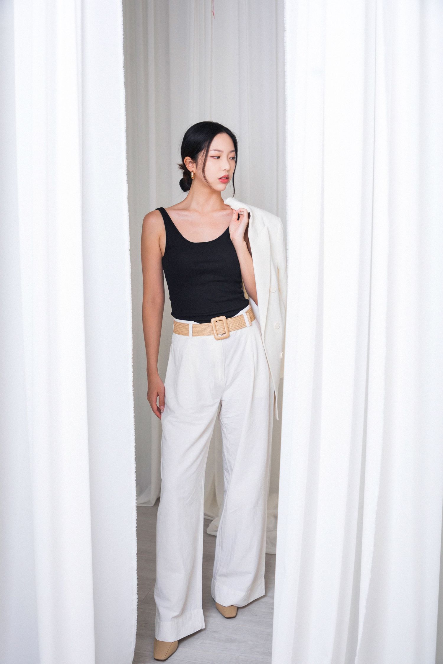 Woman in black tank top, white wide-leg trousers, and woven belt holds white blazer between white curtains.