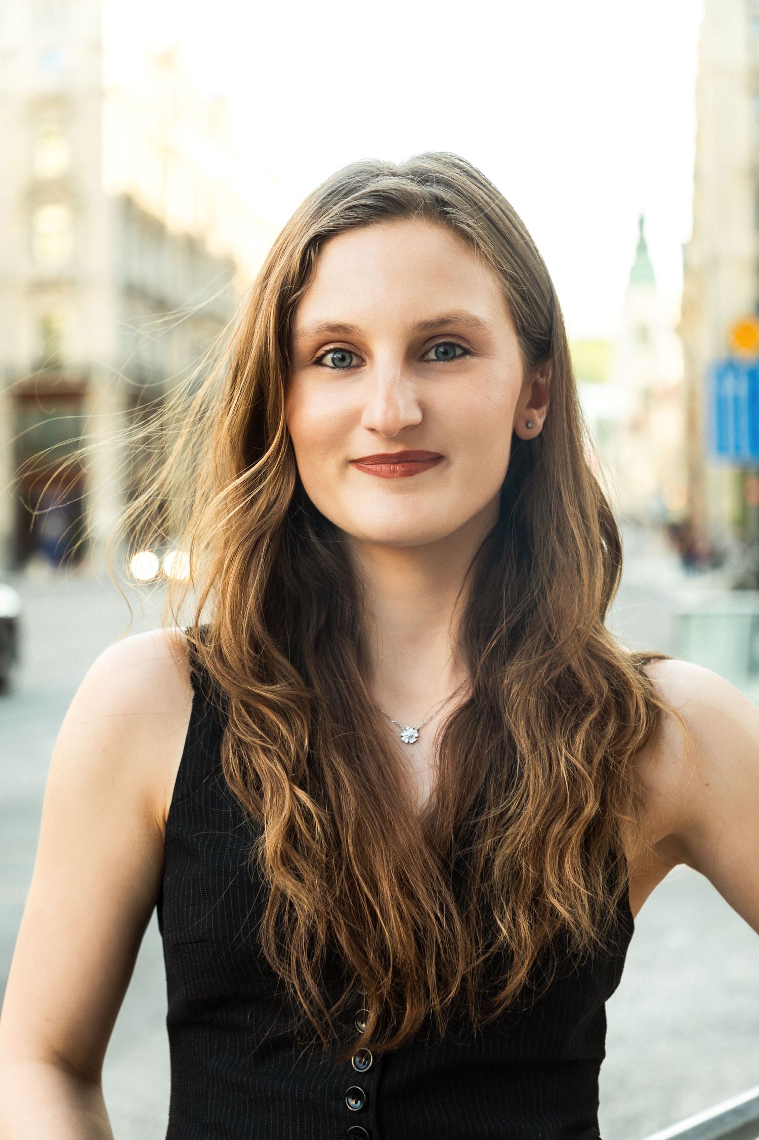 Young woman with long brown hair wearing a black sleeveless top and necklace, posing on a city street.
