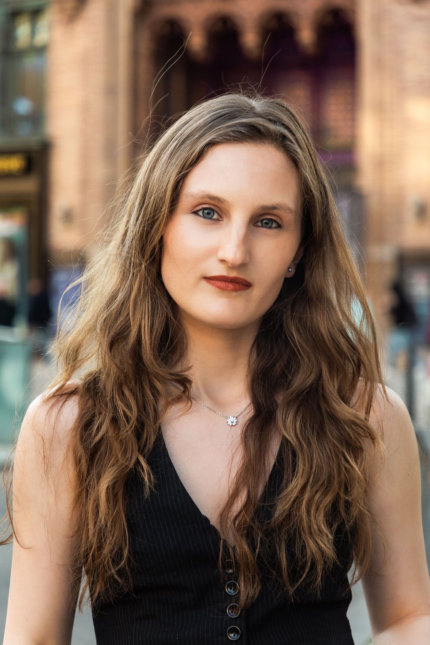 Young woman with long wavy brown hair wearing a black sleeveless top and necklace, posing outdoors in an urban setting.