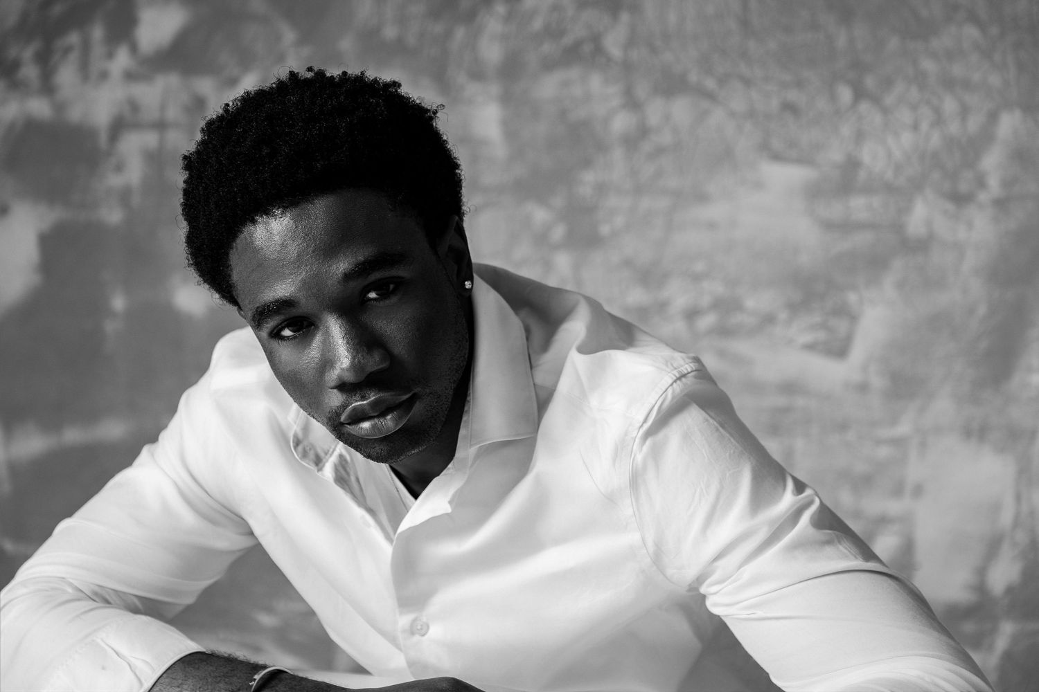 Black and white portrait of a young man in a white dress shirt, seated against a textured wall.