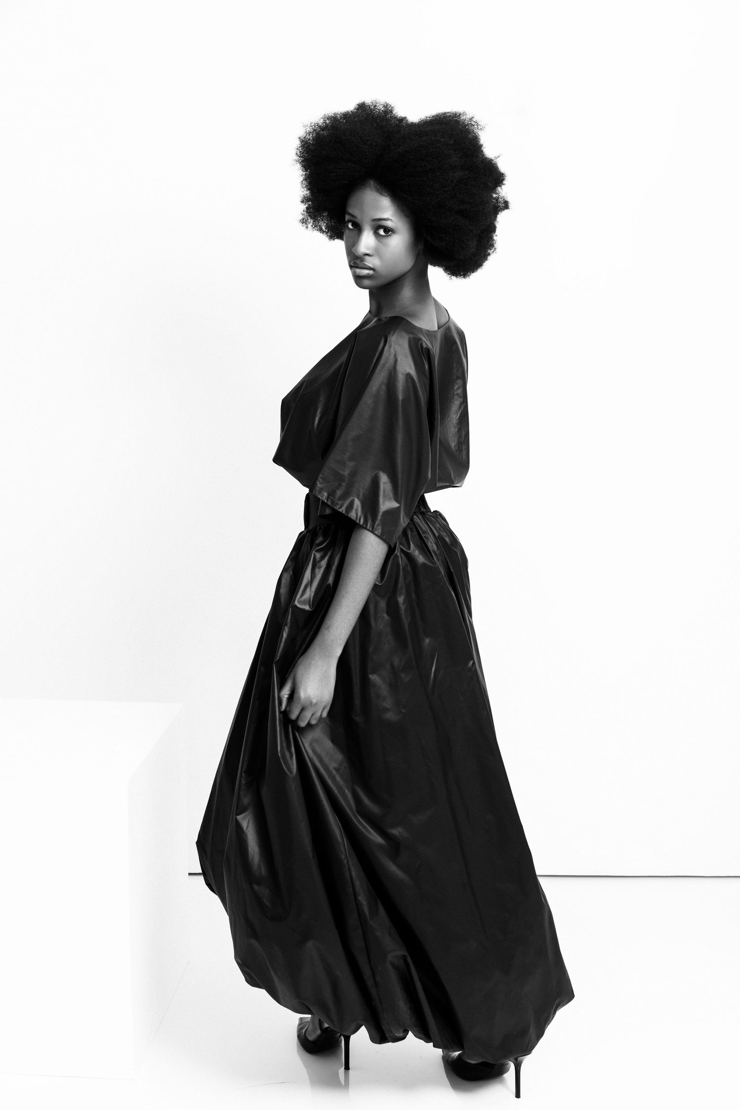 Black and white photo of a woman with an afro wearing a flowing black satin gown and heels, posing elegantly.