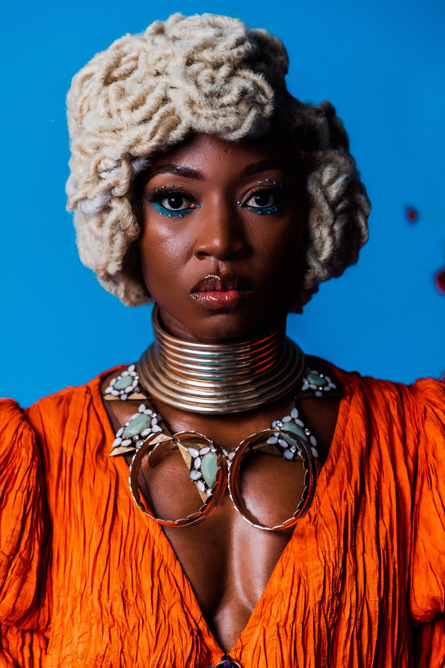 A woman in an orange dress wearing a cream knit hat, bold blue eye makeup, and layered gold neck rings poses against a blue background.