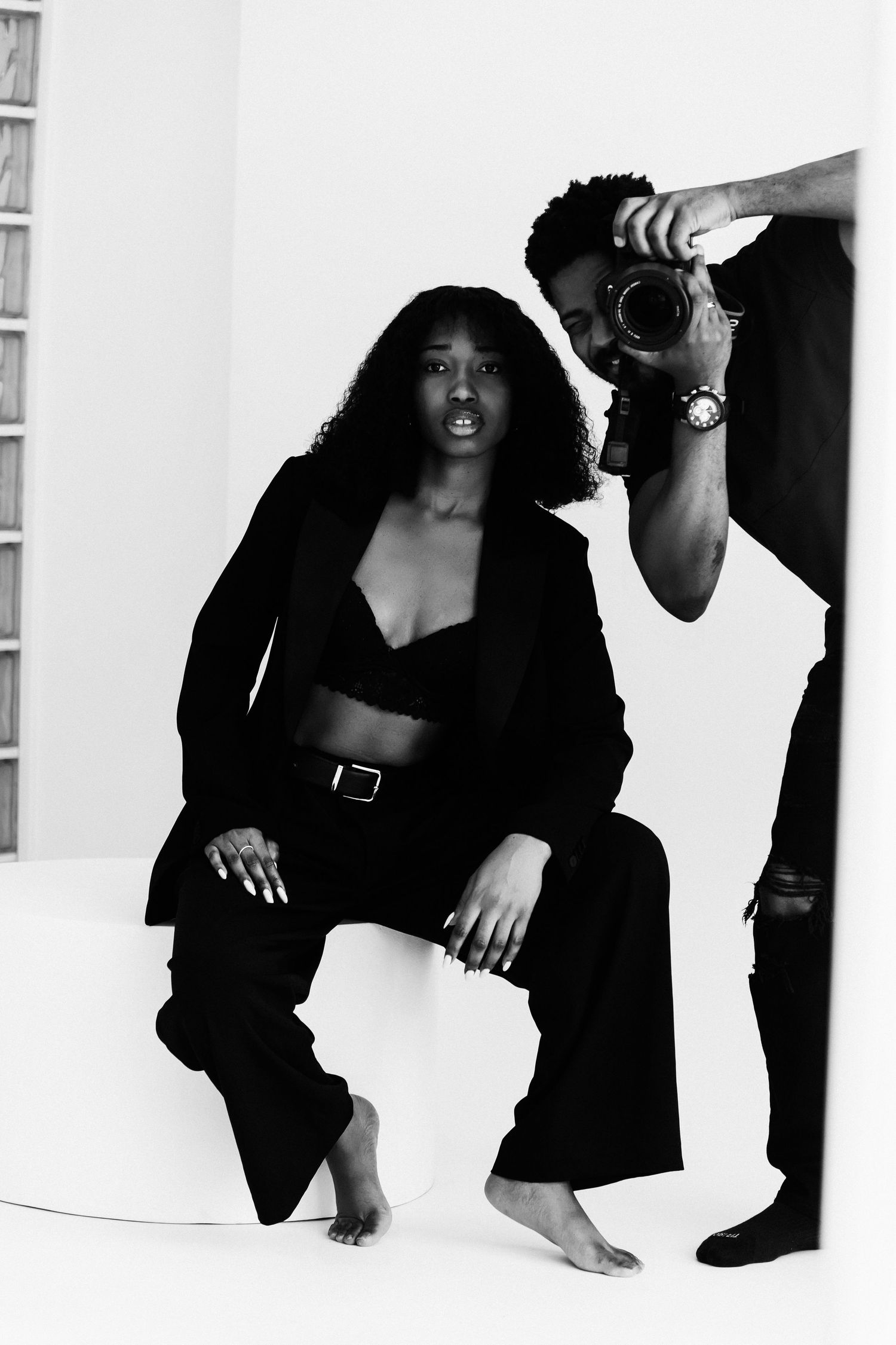 Black and white photo of a woman in black suit posing while a photographer captures her in a studio.