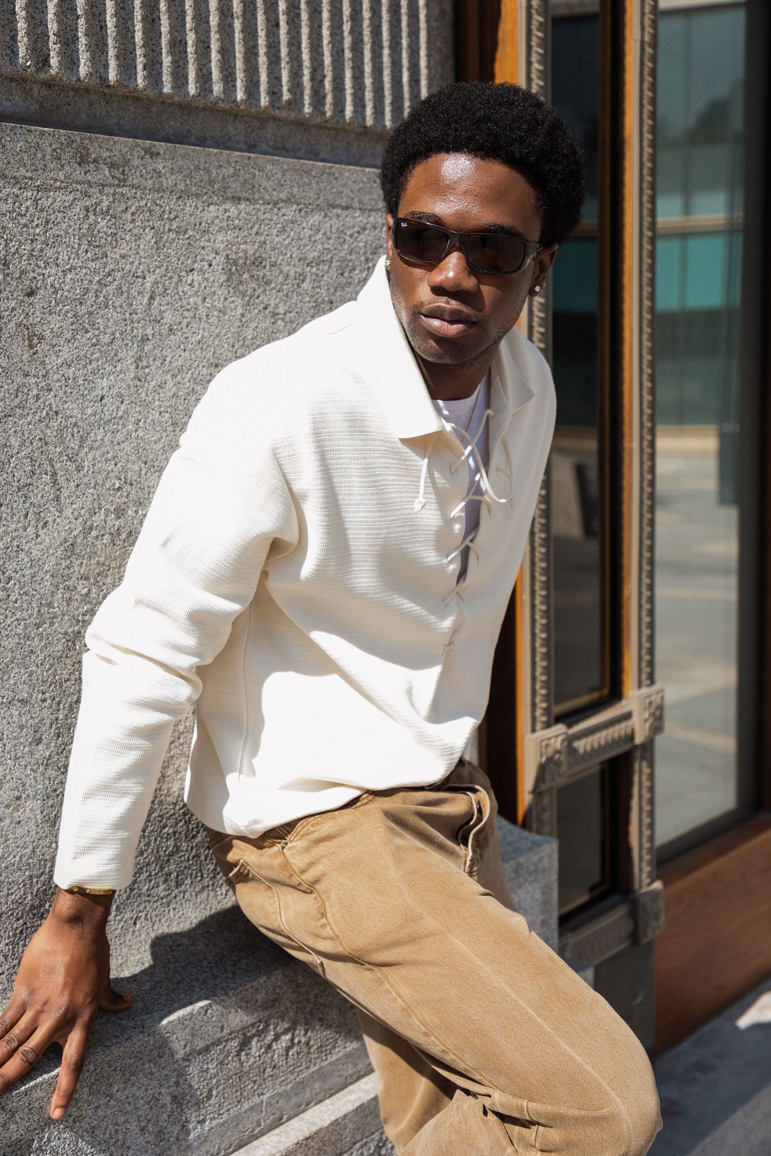Young Black man in white knit jacket and tan cargo pants leaning against a building wall wearing sunglasses.