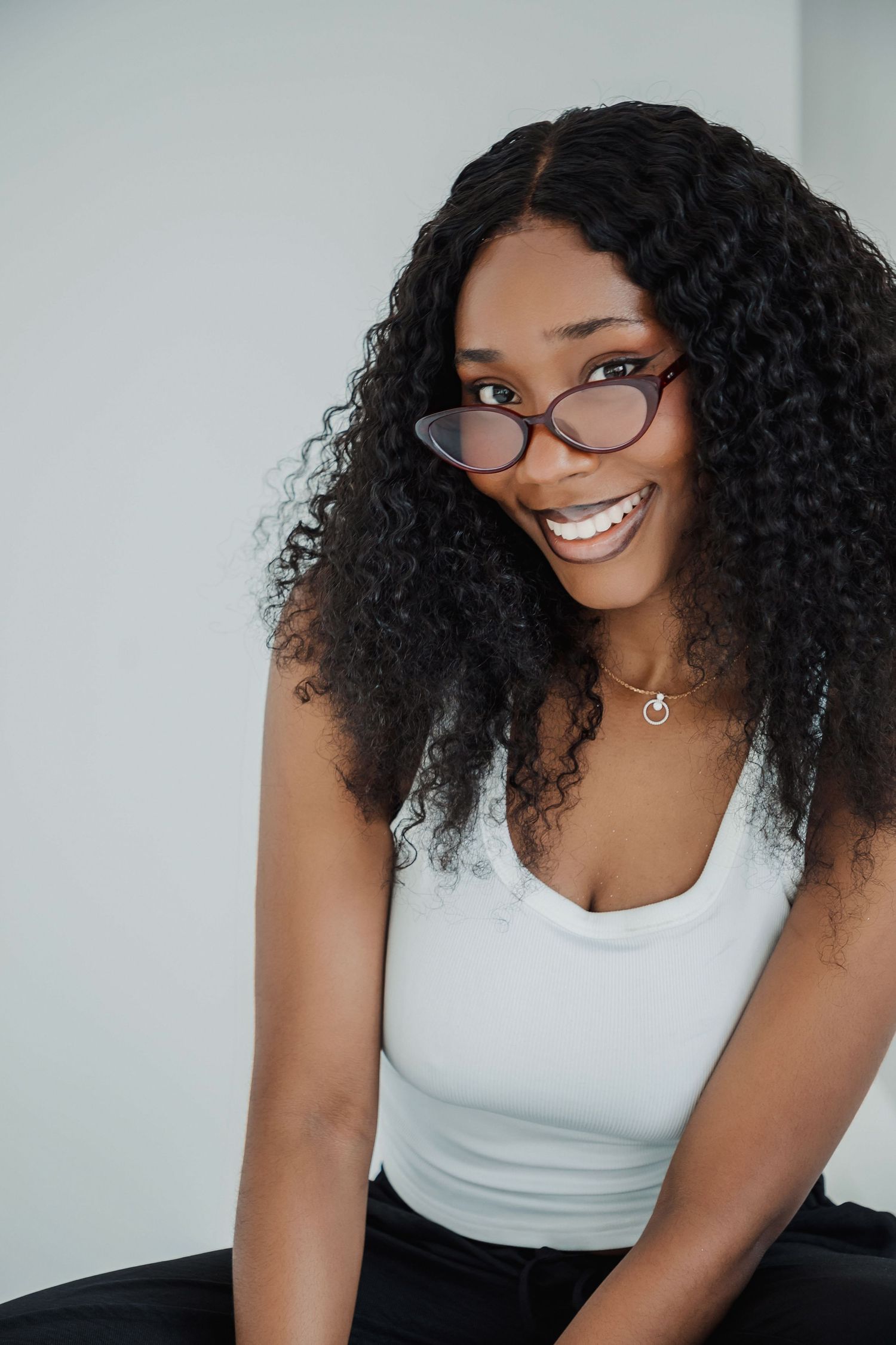 Smiling young Black woman with curly hair and cat-eye glasses wearing a white tank top against a light background.