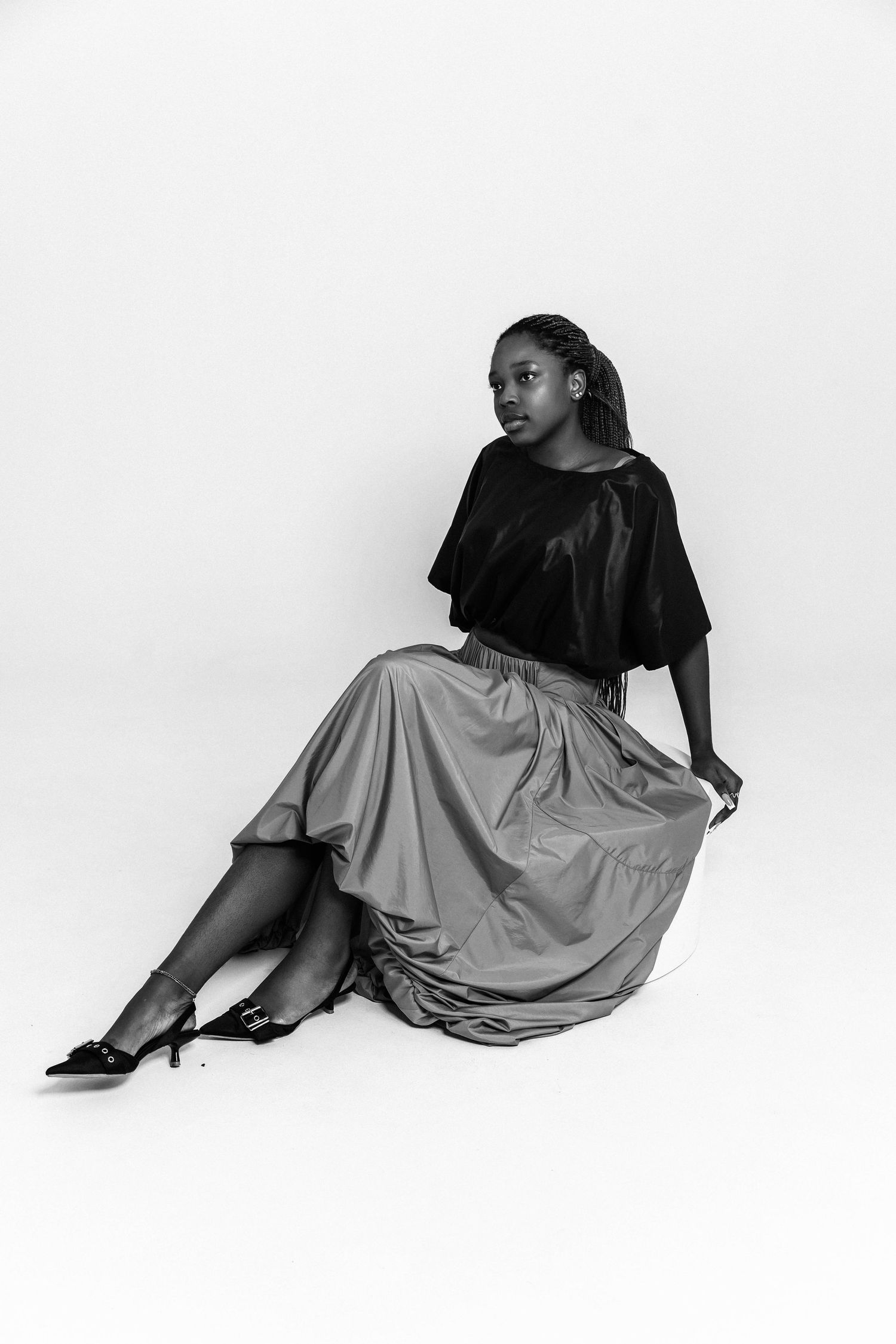 Black and white photo of a young woman in a black top and flowing midi skirt, seated gracefully in heels.