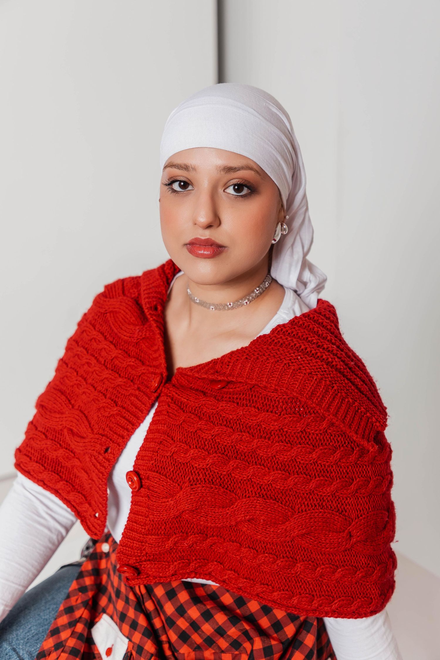 Young woman wearing white hijab and red cable-knit shawl over white top, posing indoors with silver necklace.