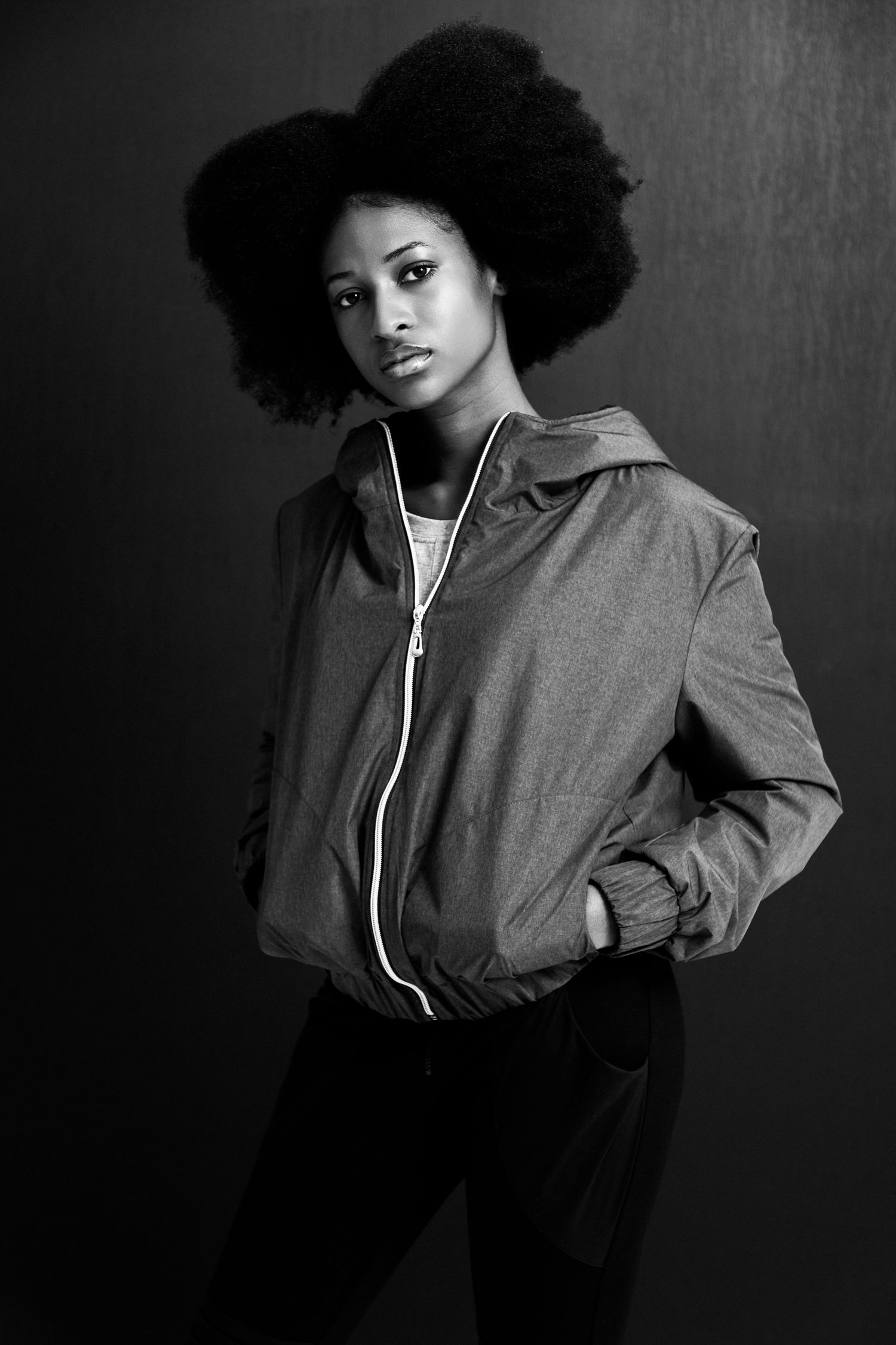 Black and white portrait of a young woman with an afro hairstyle wearing a zip-up jacket, posing confidently.
