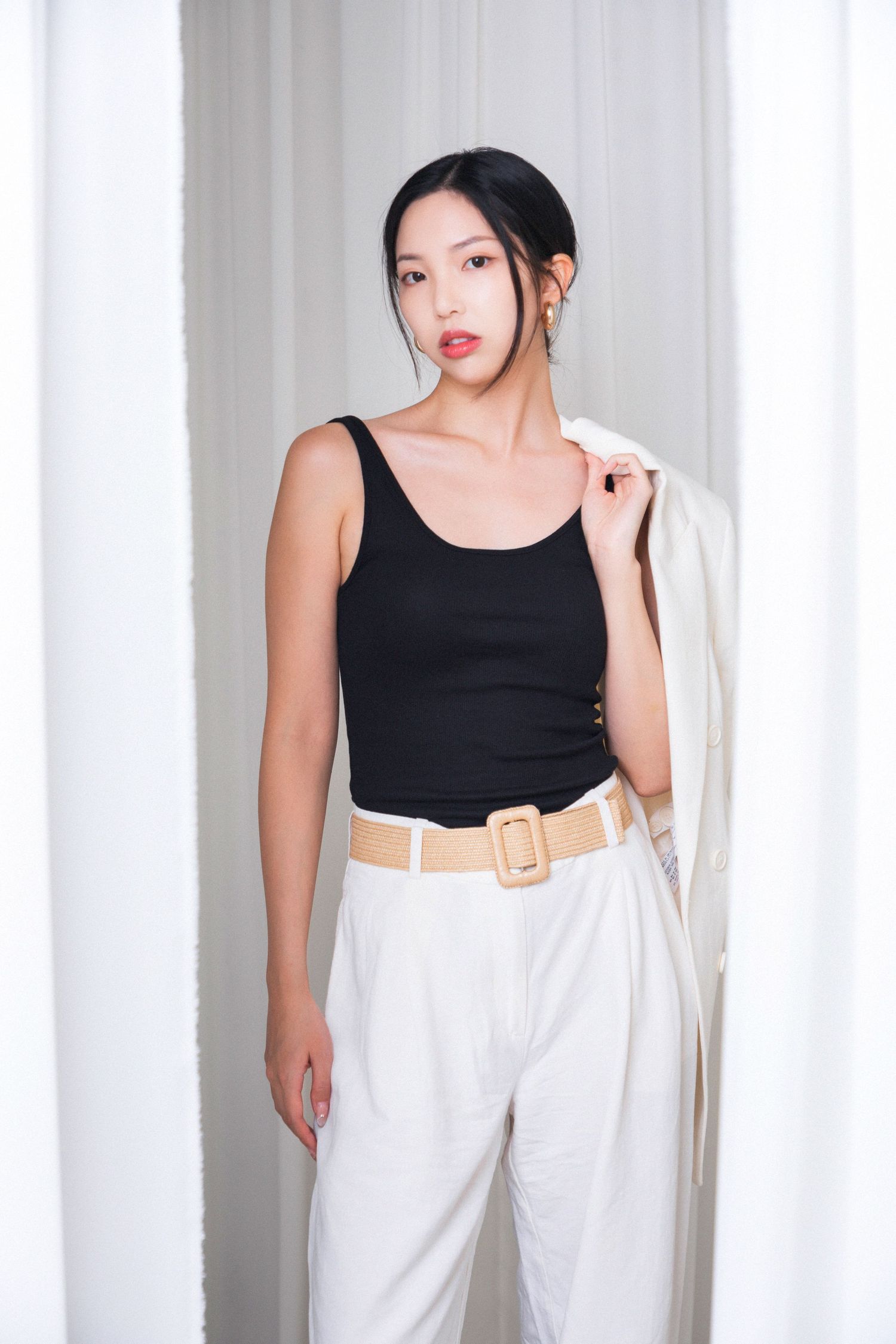 Young woman in black tank top, white trousers, and tan belt, holding a white blazer over her shoulder.