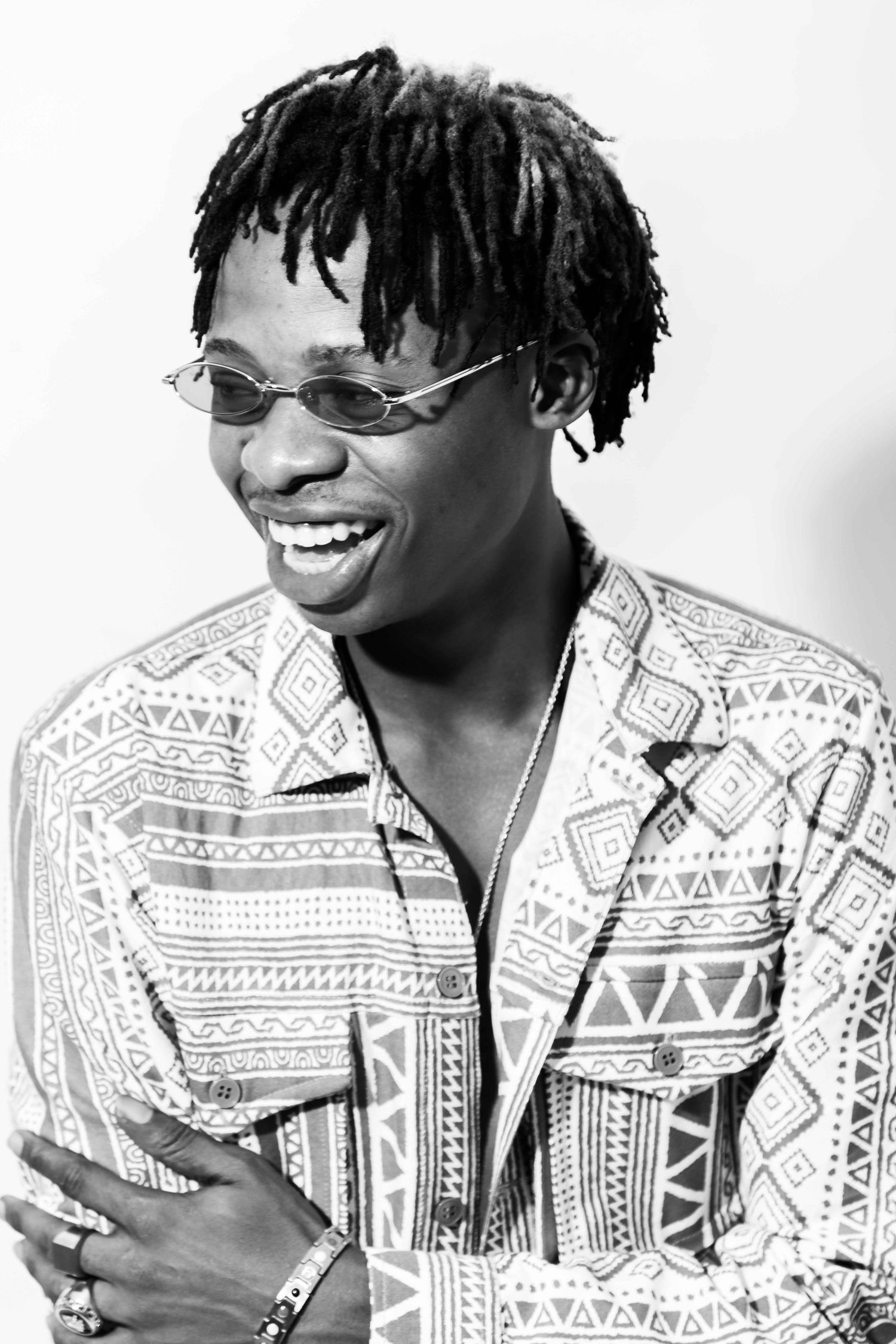 Black and white portrait of a smiling young man with dreadlocks, oval sunglasses, and a patterned shirt.