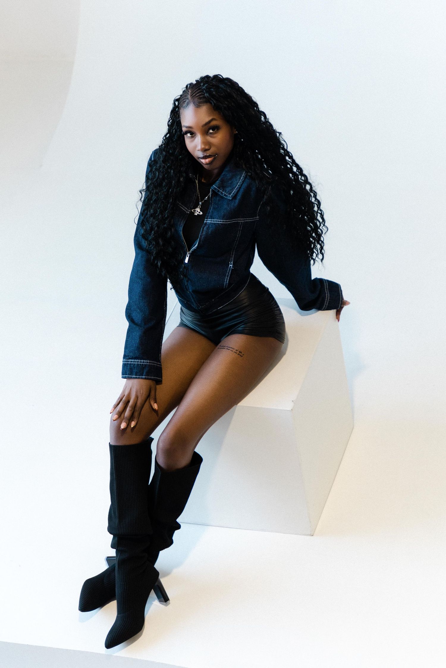 A young Black woman with long curly hair poses in a dark denim outfit and black knee-high boots on a white cube.