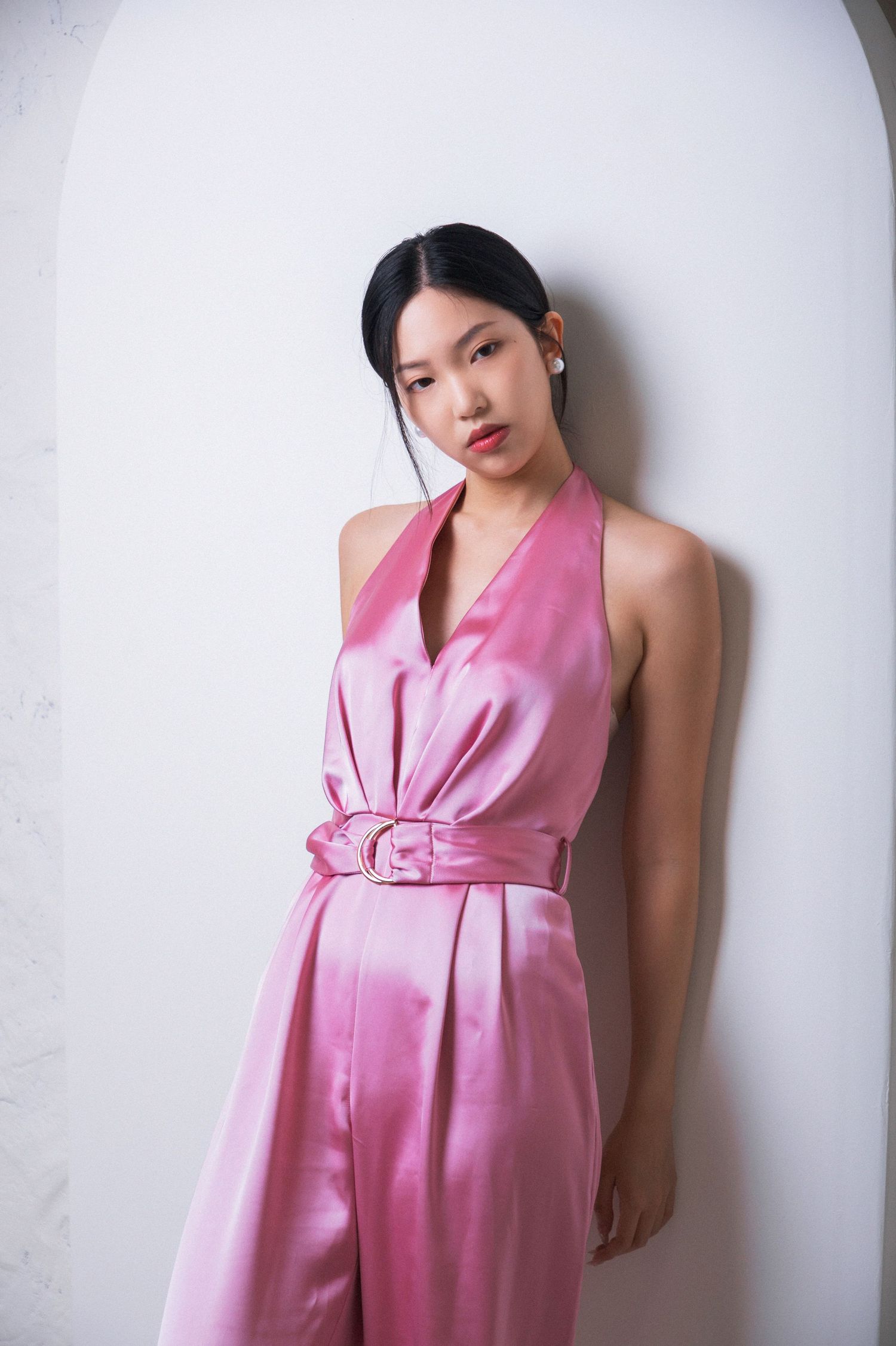 Young Asian woman posing in a pink satin halter jumpsuit with belt against a white wall.