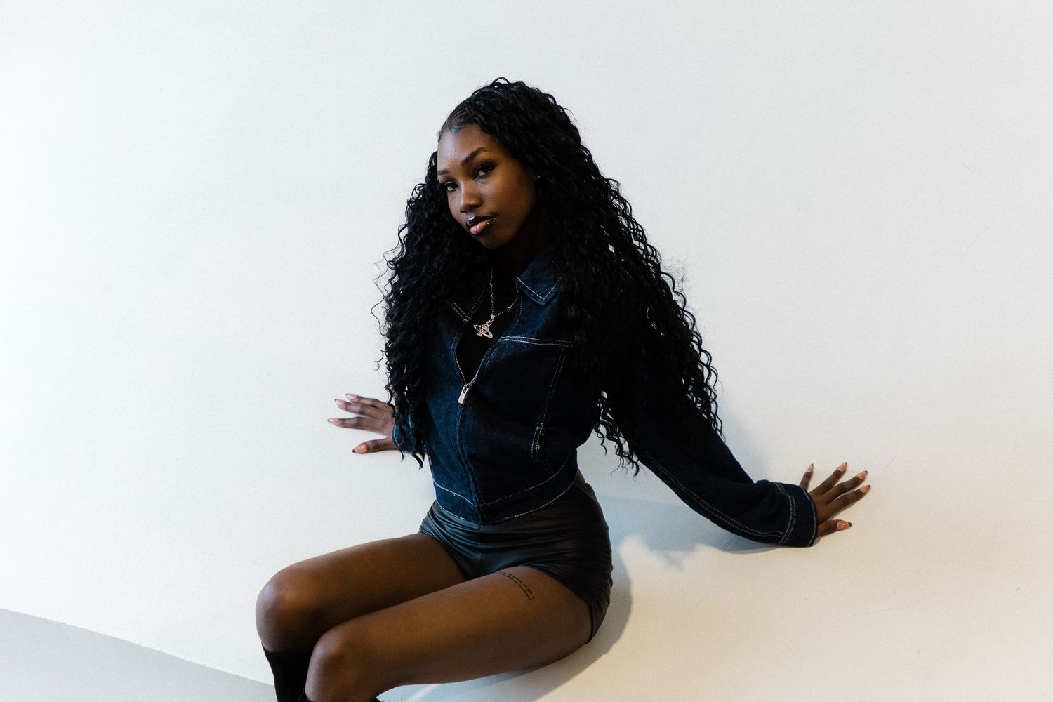 Young Black woman with long curly hair poses confidently in a black mini dress against a white wall.