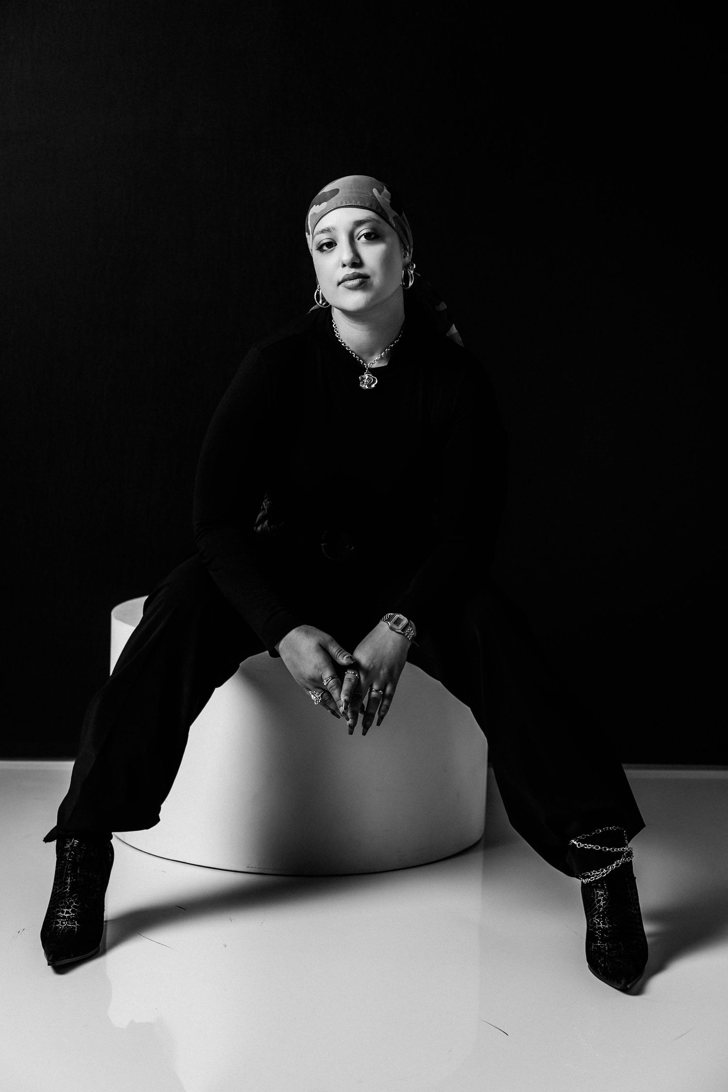 Black and white portrait of a woman in dark clothing and head wrap sitting on a white stool against black background.