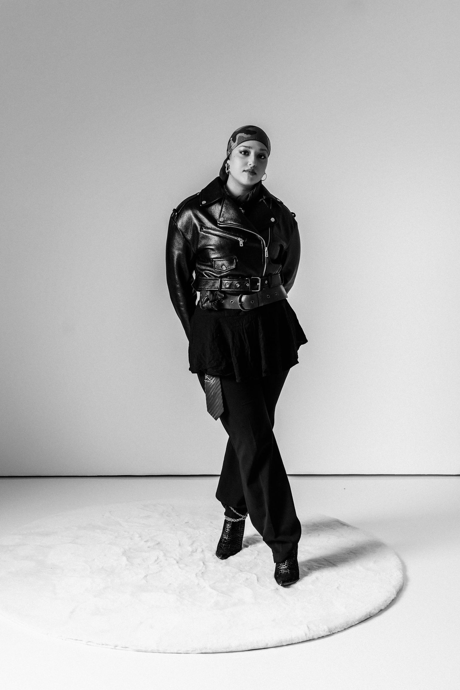 Person in leather biker jacket, black wide-leg pants, and boots standing on round white rug in black and white photo.