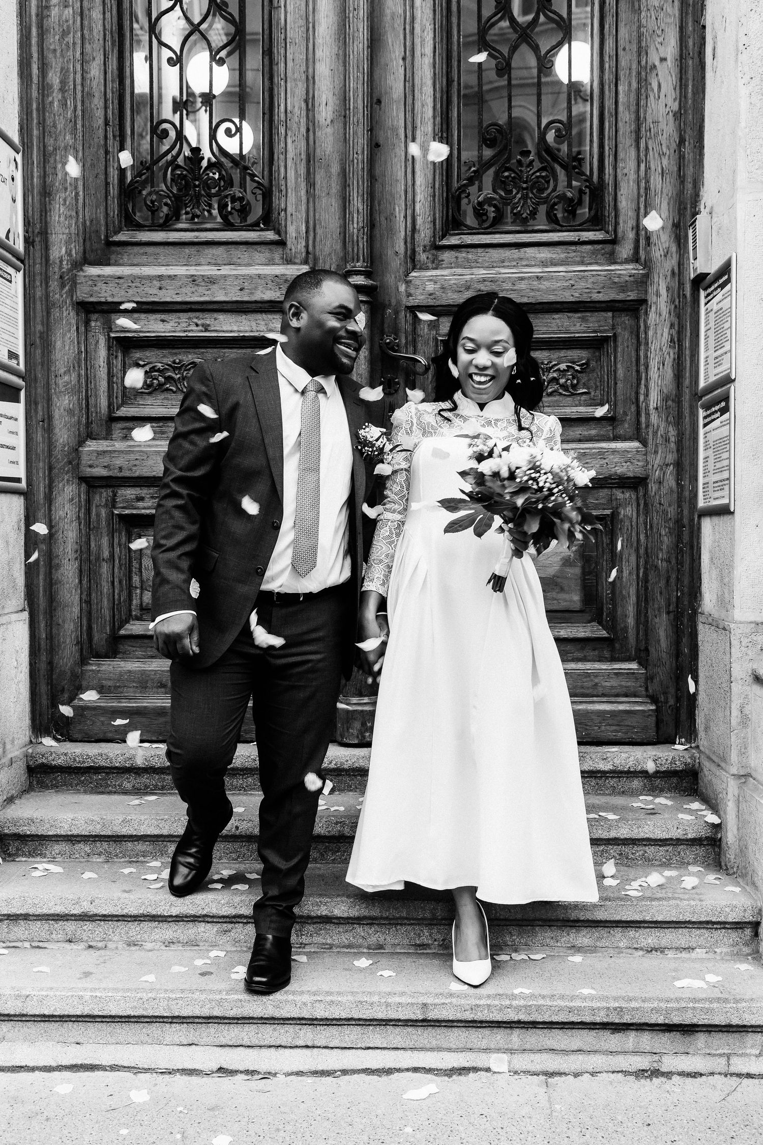 Black and white photo of a joyful newlywed couple descending church steps with confetti falling around them.