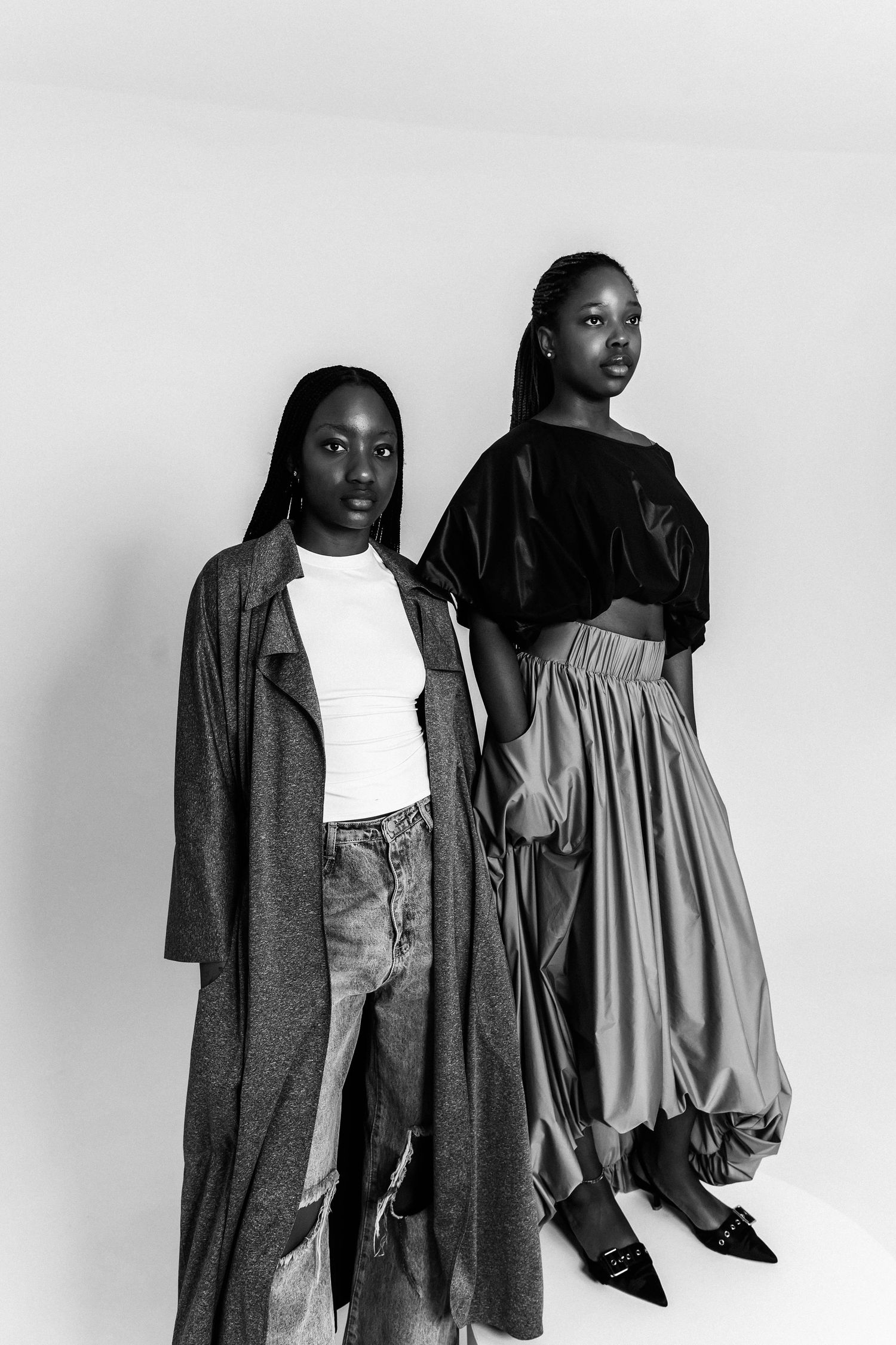 Two Black women pose in stylish monochrome outfits, one in a long coat with jeans, the other in a velvet top and flowing skirt.