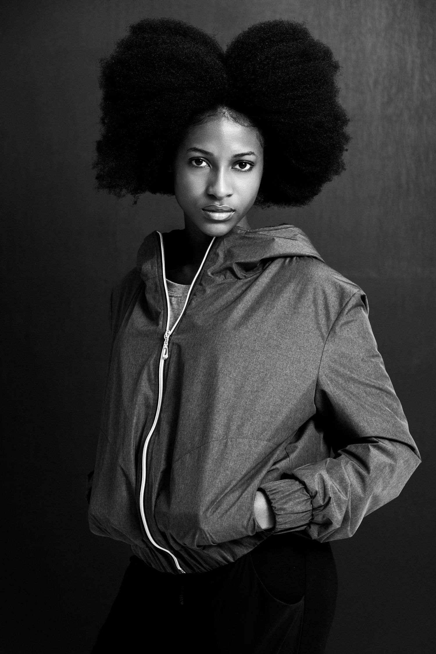 Black and white portrait of a young woman with a large afro hairstyle wearing a zip-up hoodie jacket.