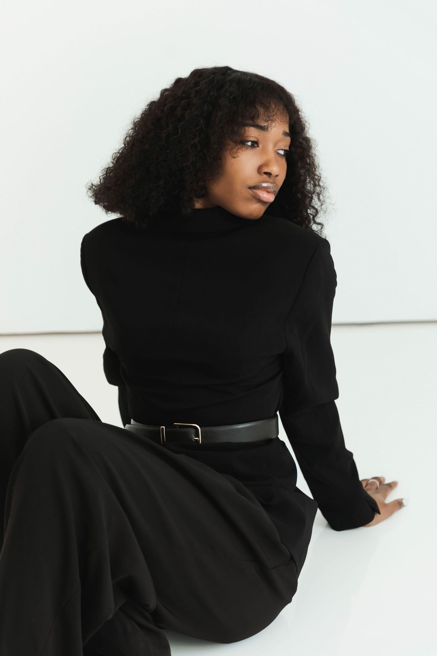 Woman in black turtleneck and wide-leg pants sitting on white floor, looking over shoulder with curly hair.