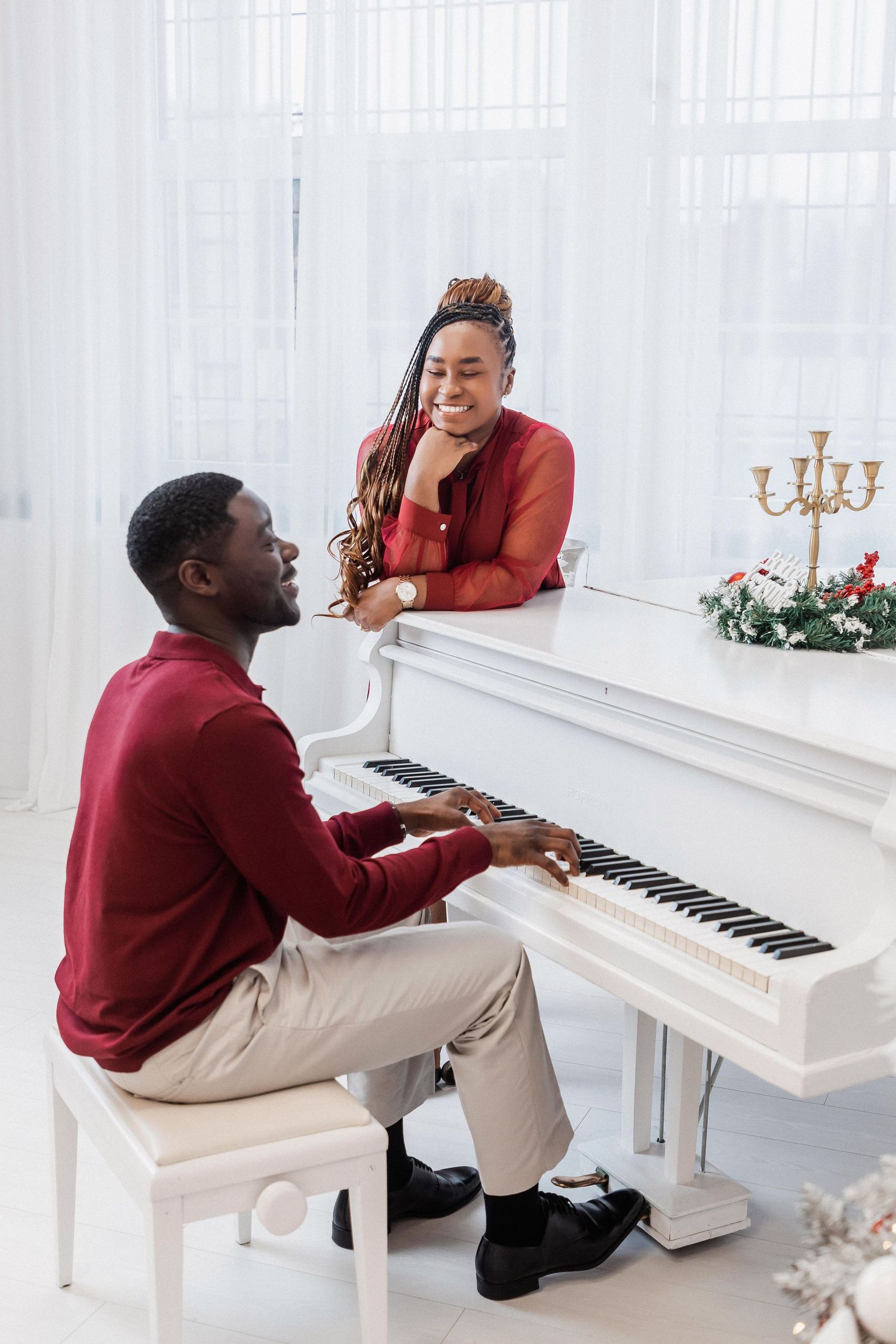 A man plays white grand piano while a smiling woman leans on it, both wearing red in a bright white room.