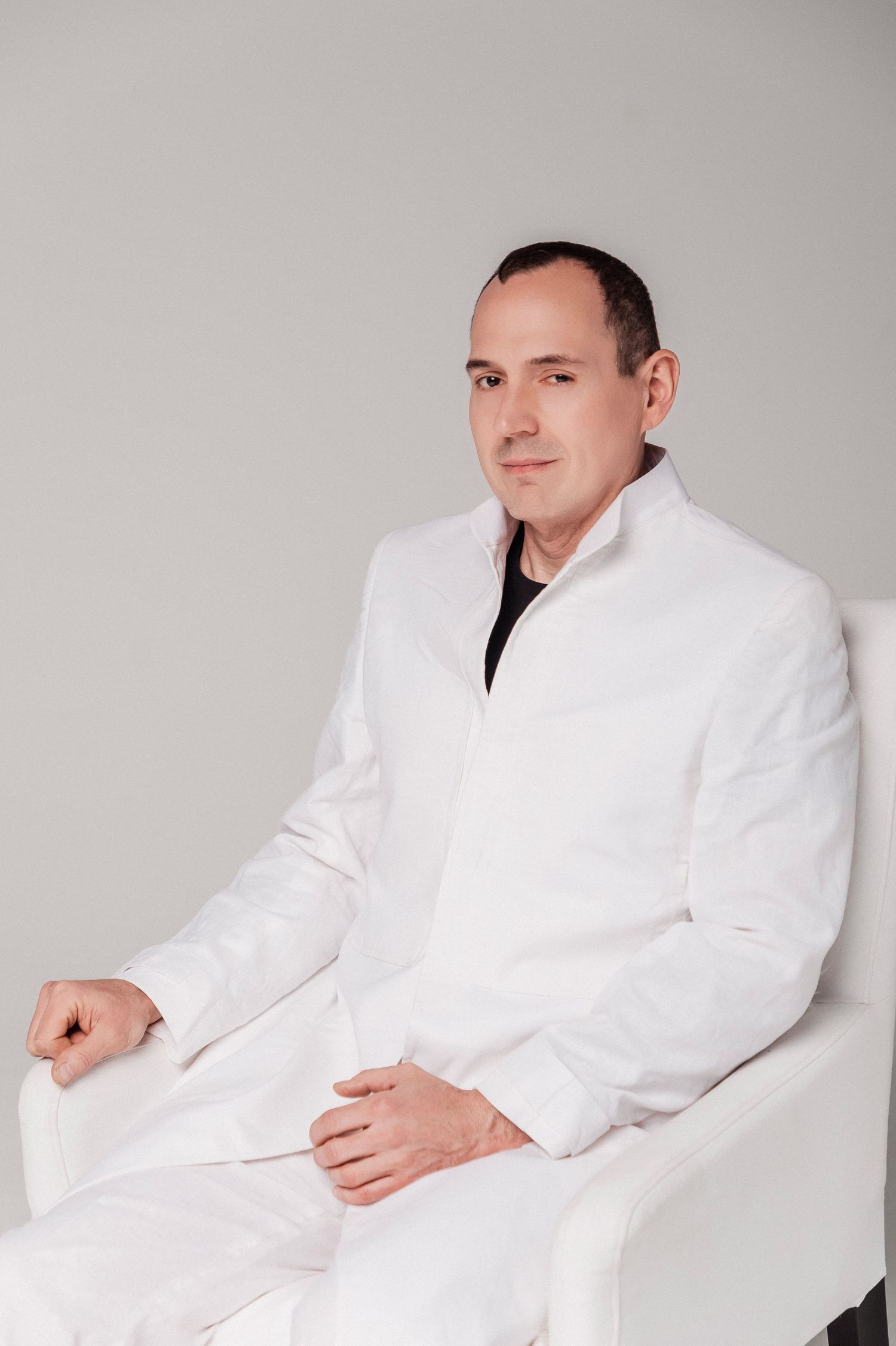 A man in a white medical coat sits relaxed in a white chair against a light gray background.