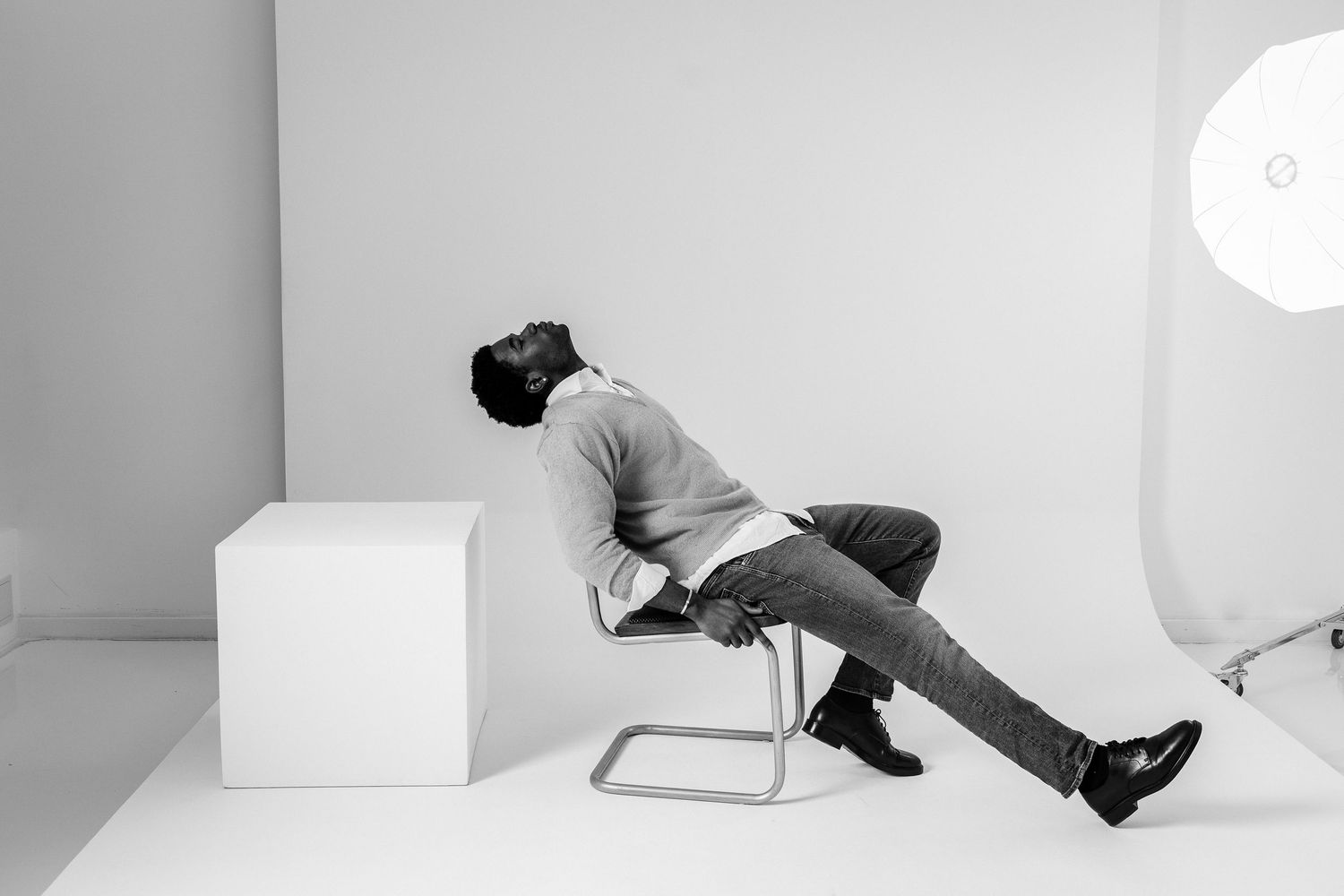 Black and white photo of a man leaning back relaxed on a chair in a minimalist photography studio setting.