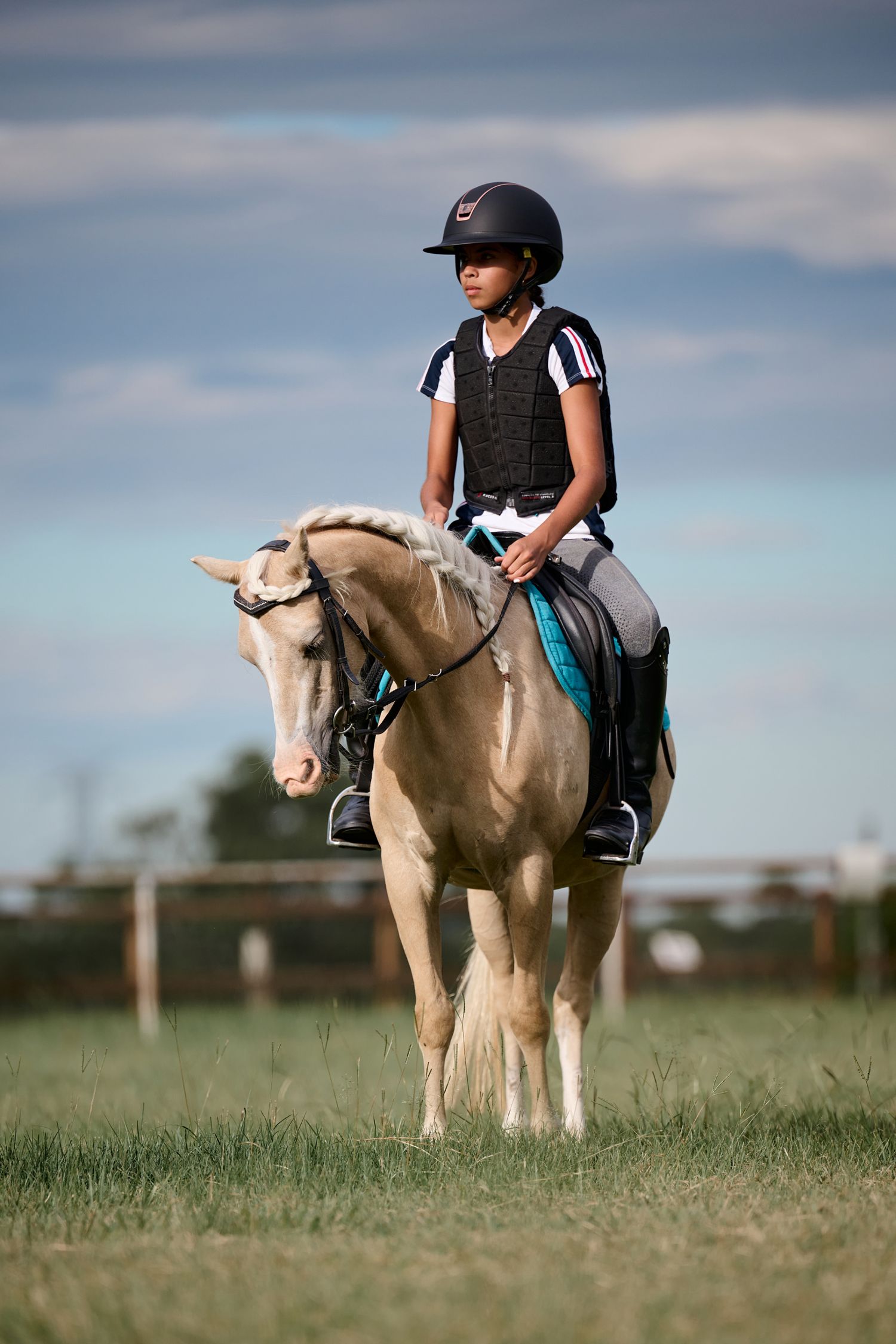 An equestrian rider wearing safety gear sits atop a pale cream-colored horse in a grassy field under cloudy skies.