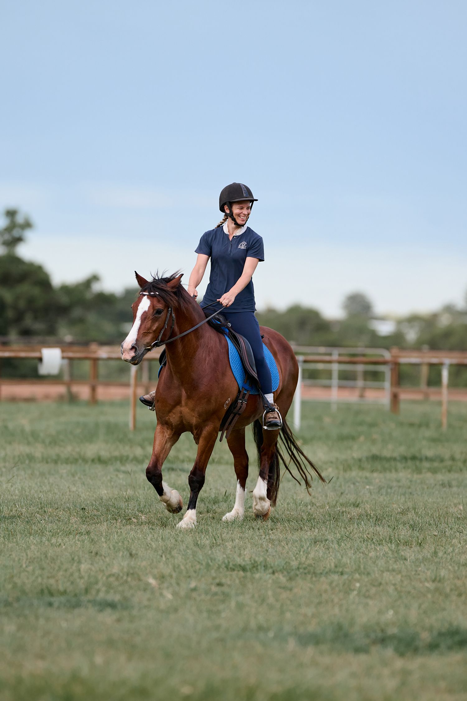 Equestrian rider in navy shirt and helmet riding brown horse in fenced pasture on sunny day.