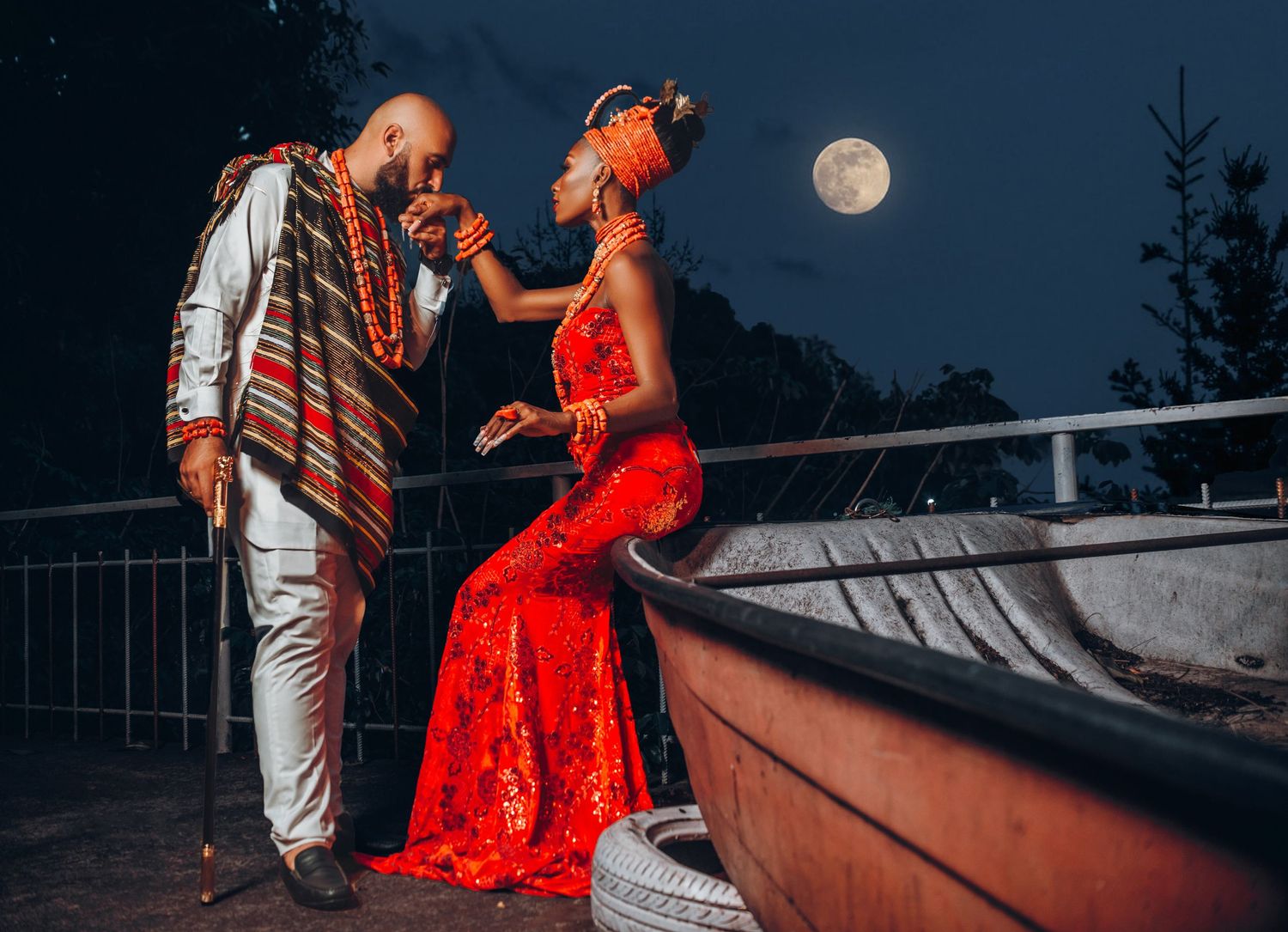 A pre-wedding photo of a couple, with the lady seated on a boat as the man gently kisses her hand, under a glowing moon.