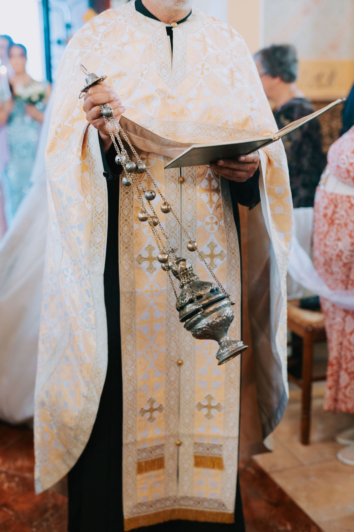 Orthodox priest wearing white embroidered vestments holds a ceremonial censer during religious service.
