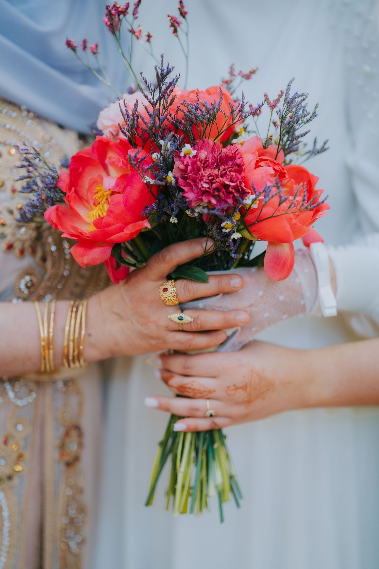 Hands adorned with gold jewelry hold a vibrant coral peony bouquet with purple accents against a soft blue backdrop.