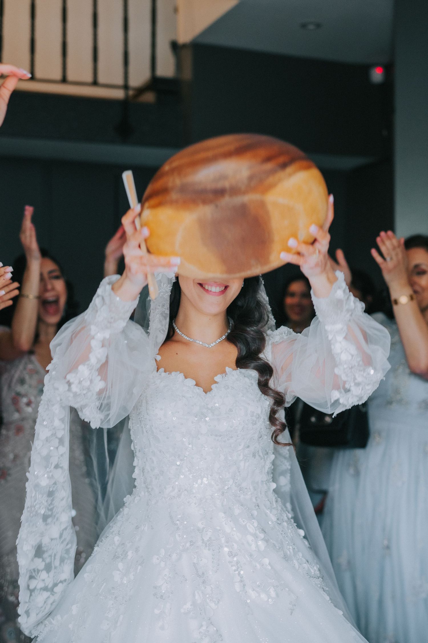 Bride in white lace wedding dress playfully holds up round piece of bread during wedding celebration.