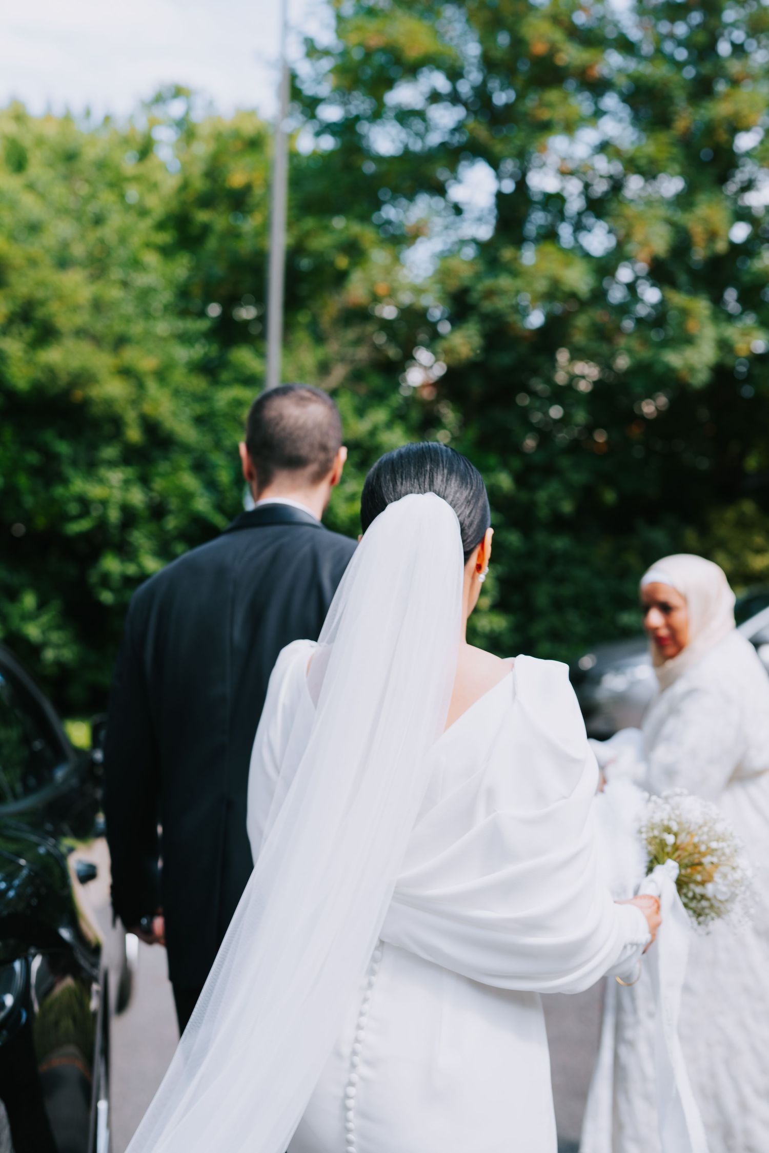 Bride in flowing white dress with veil standing outdoors during wedding ceremony surrounded by green trees.