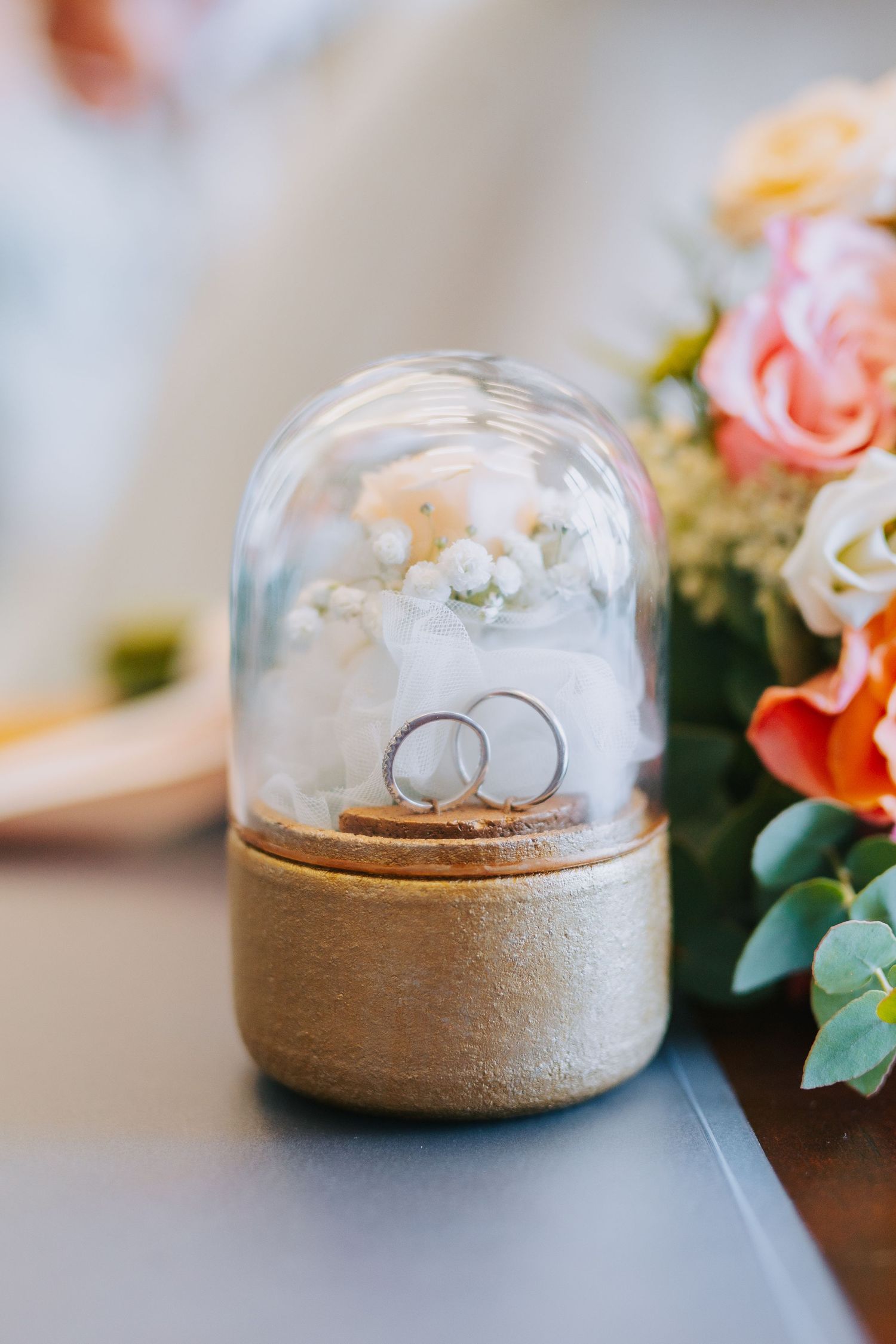 Wedding rings displayed in a small glass cloche dome with floral decorations beside it on a wooden surface.