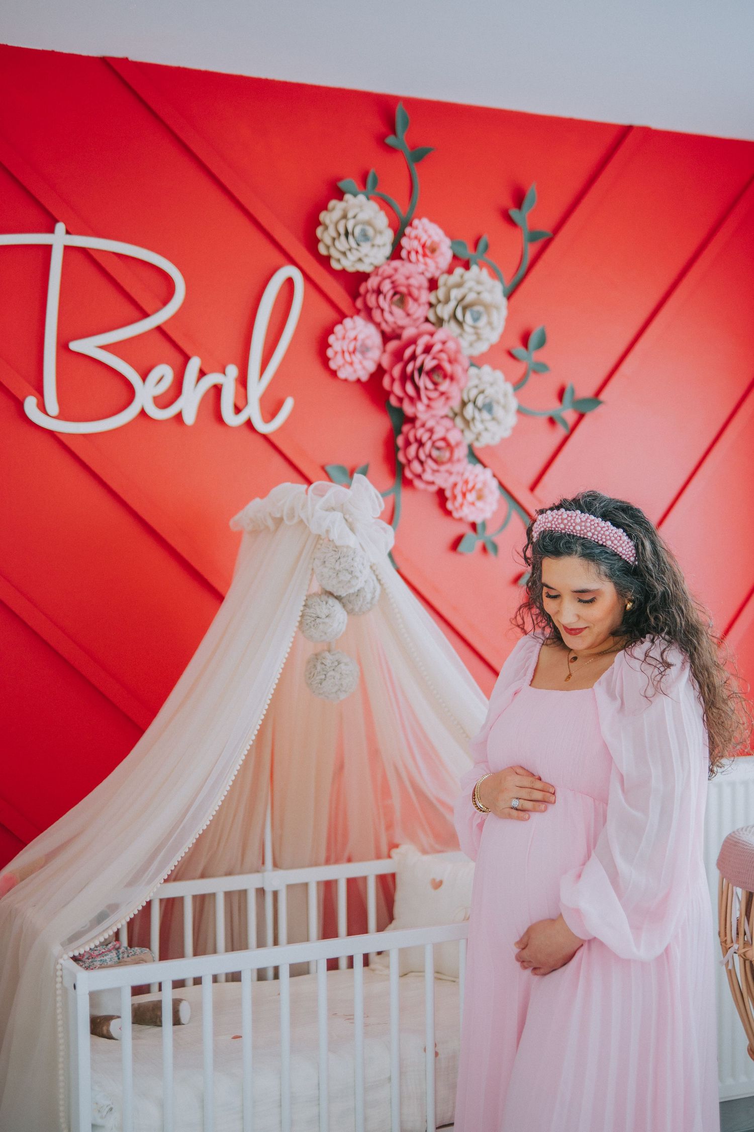 Maternity photo with red accent wall, white crib, and floral decorations.