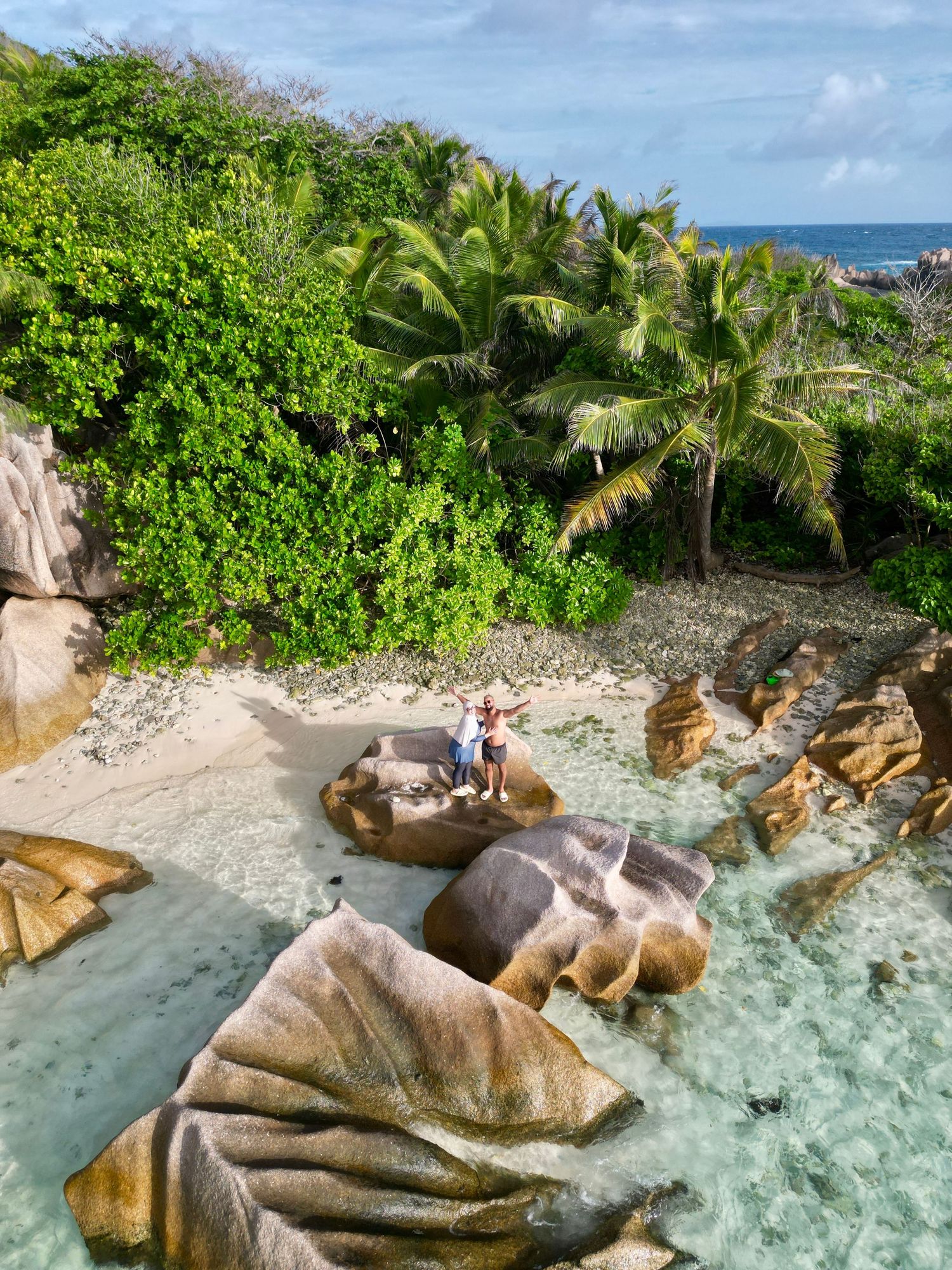 Tropical beach cove with large granite boulders, palm trees, and clear turquoise waters.