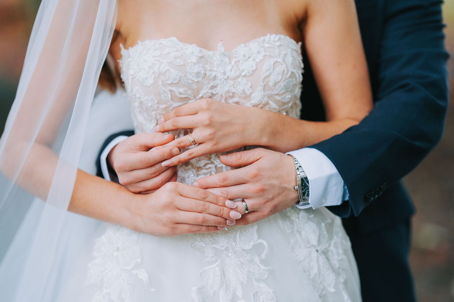 Close-up of bride and groom's hands embracing, showing wedding rings against her white lace dress and his formal suit.