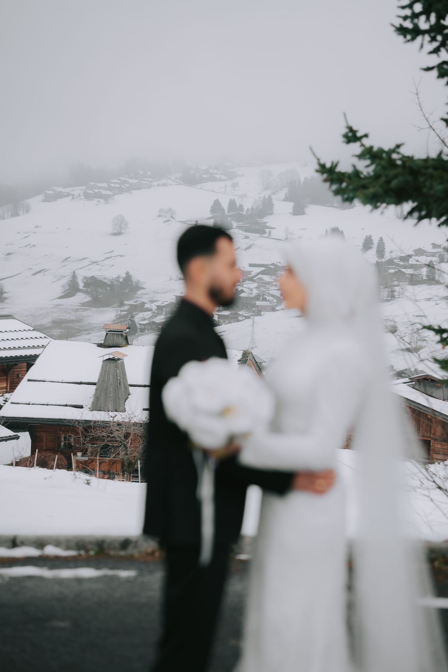 Winter wedding couple standing on snowy mountain overlook with snow-covered chalets and misty peaks in background.