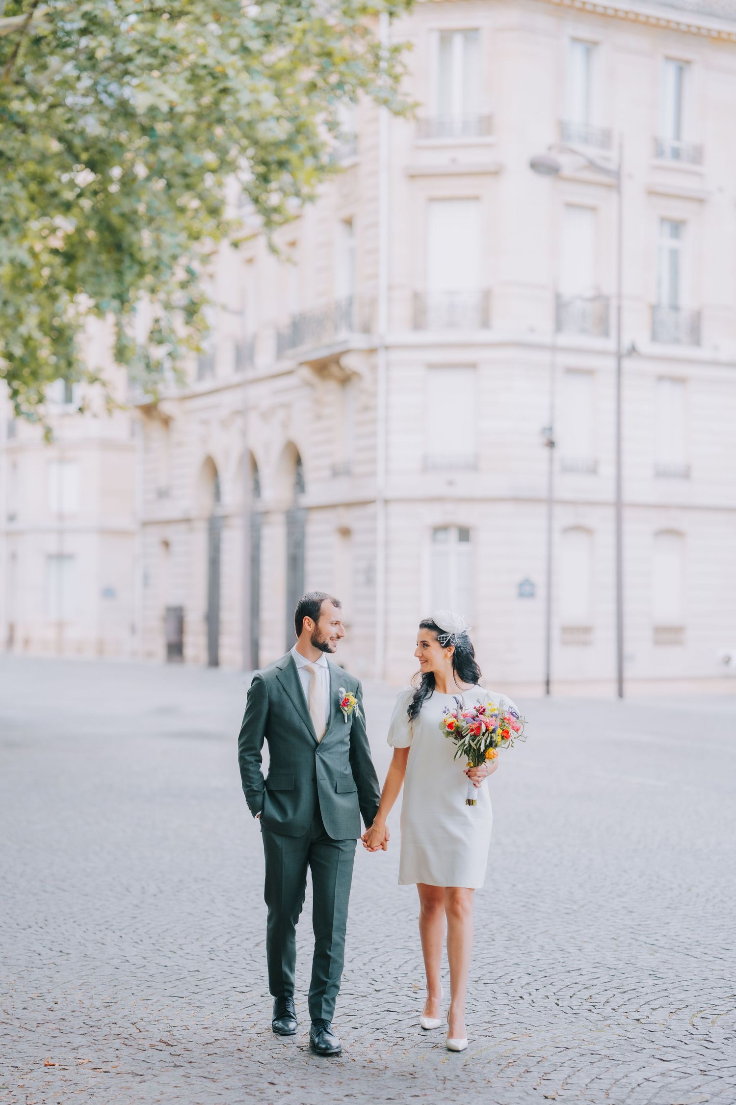 Couple walking on cobblestone street beside elegant European building, bride in short white dress holding colorful bouquet.