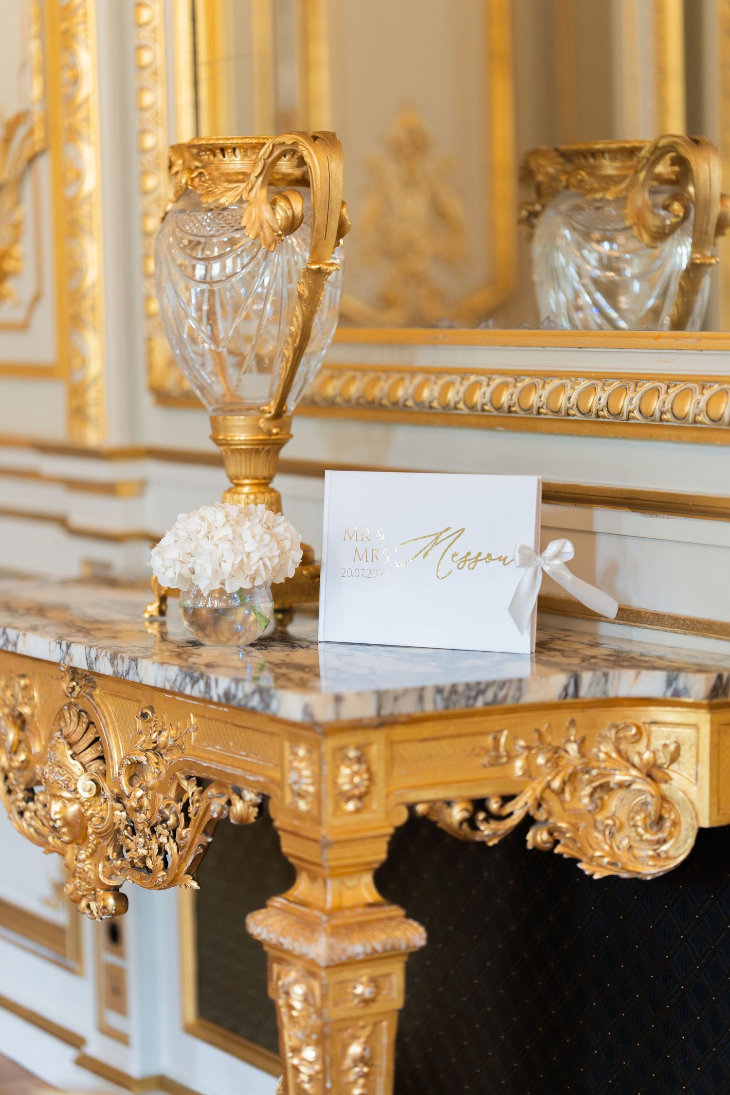 Elegant white wedding place card sits on ornate gold table with crystal vase in luxurious French palace setting.