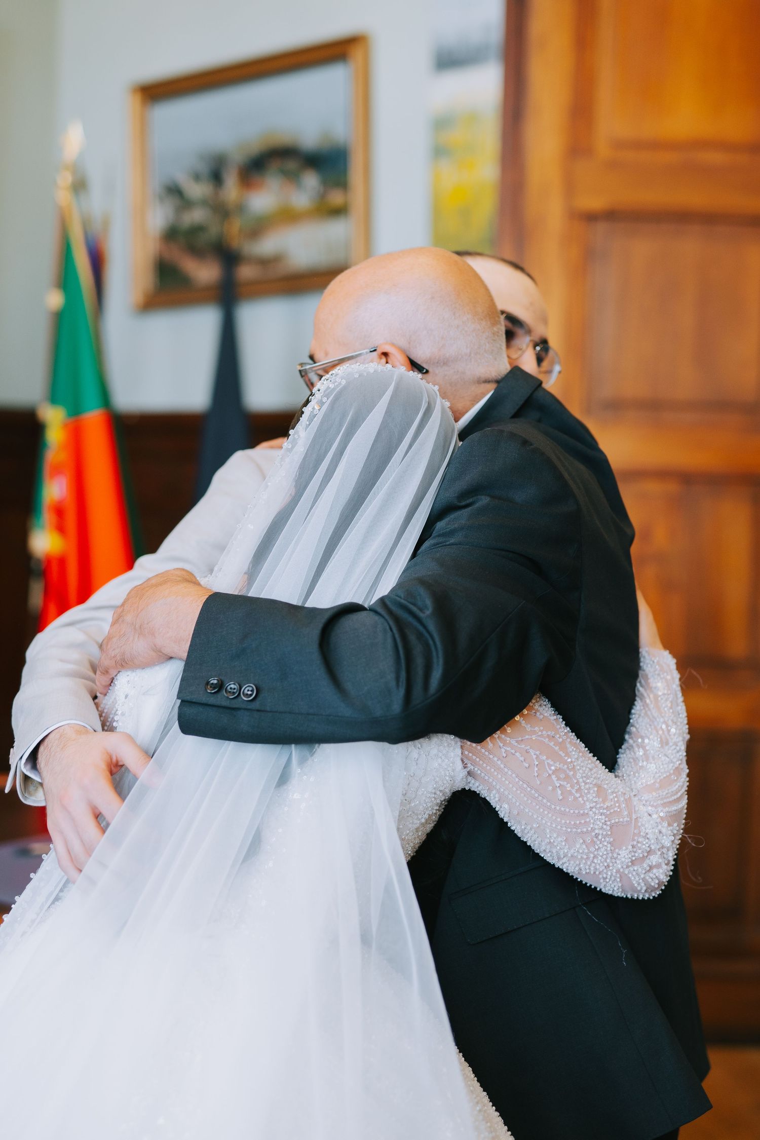 An emotional embrace between a person in a dark suit and someone wearing a white wedding dress and veil at a civil ceremony.