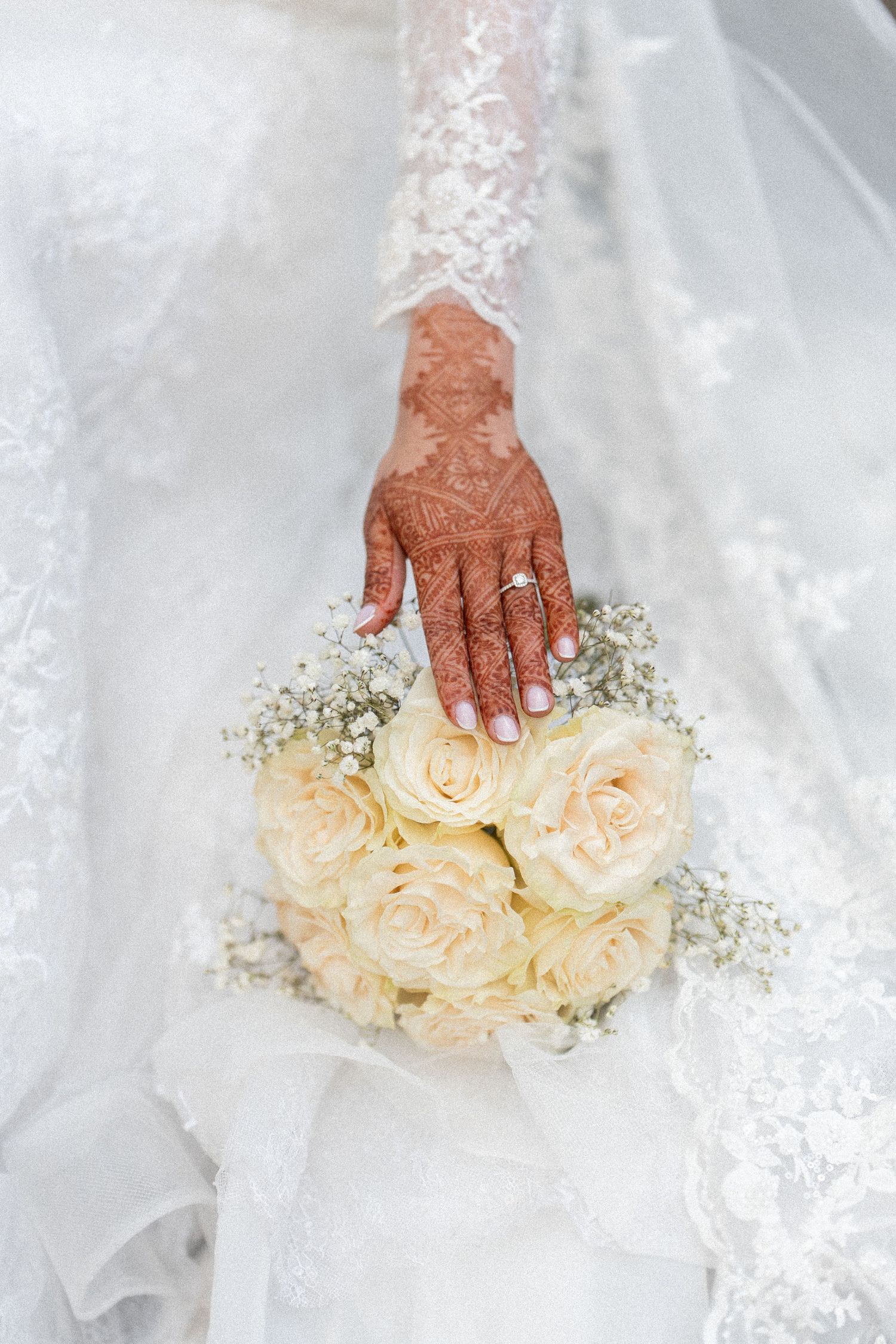 A henna-adorned hand gently touches delicate cream roses and baby's breath flowers against white tulle fabric.