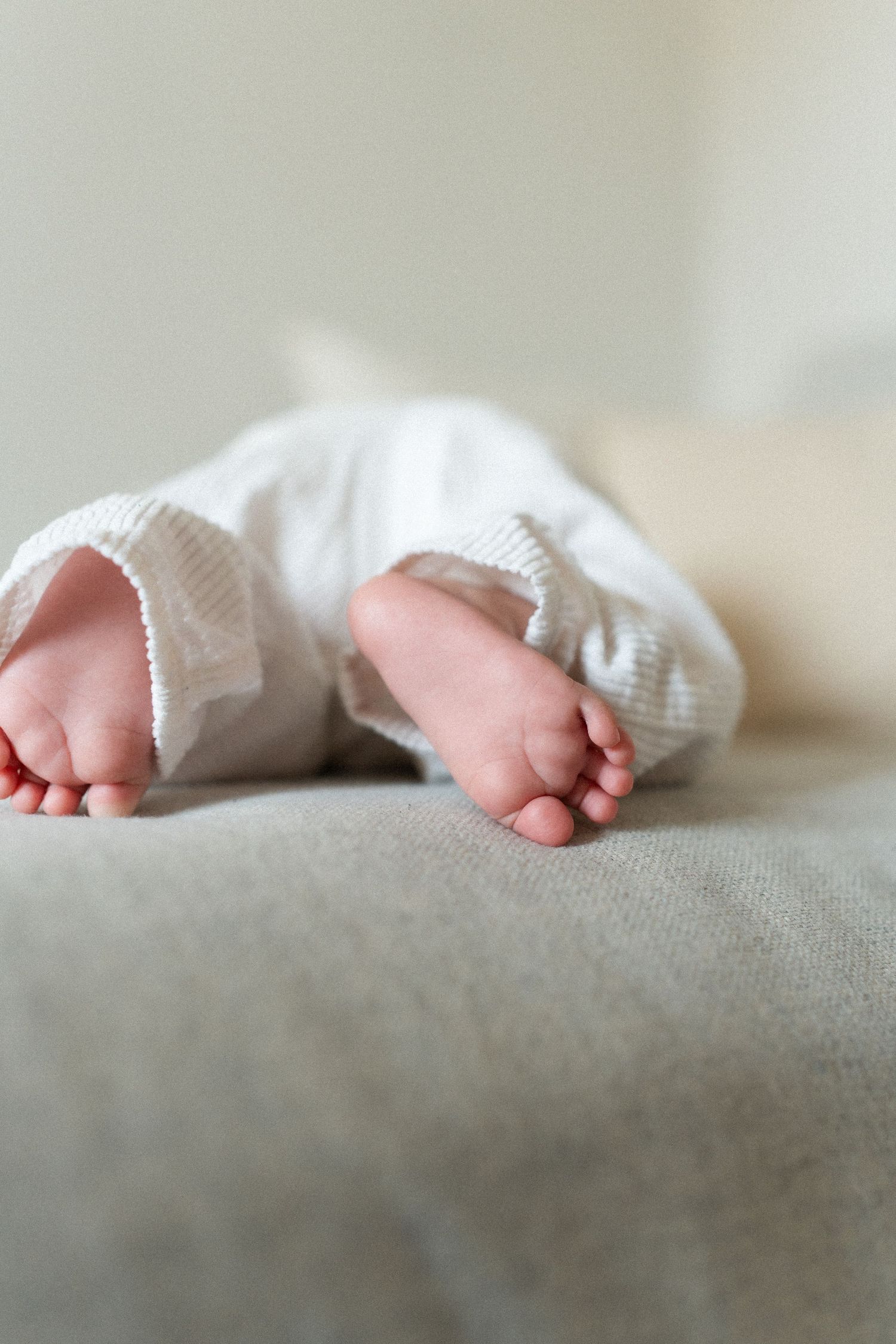 Tiny baby feet peek out from under a white knit blanket while resting on a soft gray surface.