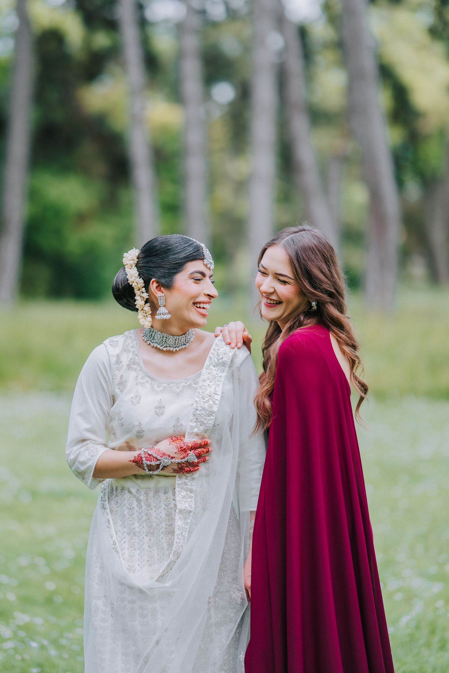 Two women in formal attire share laughter in an outdoor setting with trees in the background.