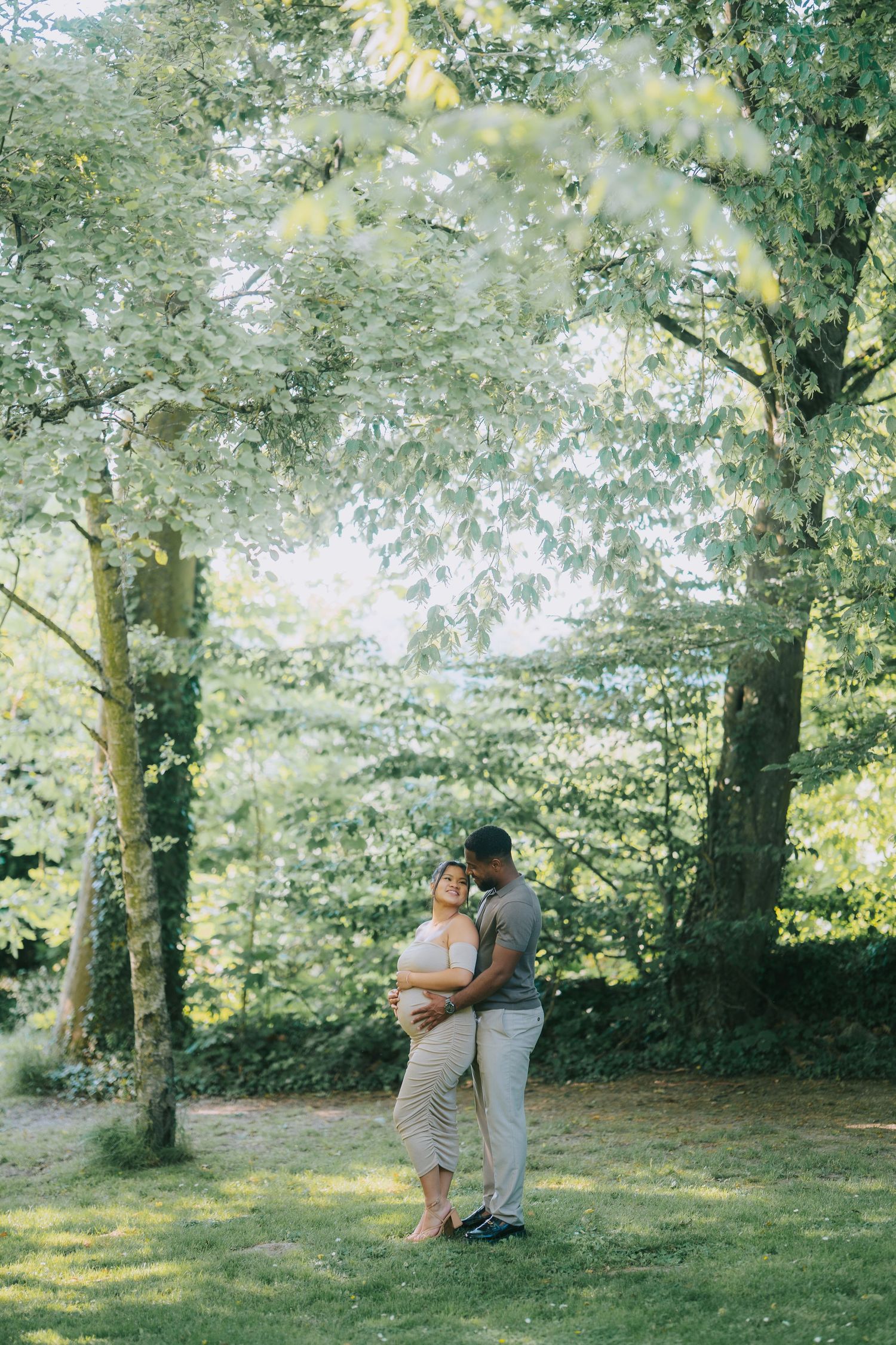A couple shares a tender embrace in a sunlit garden surrounded by lush green trees on a summer afternoon.