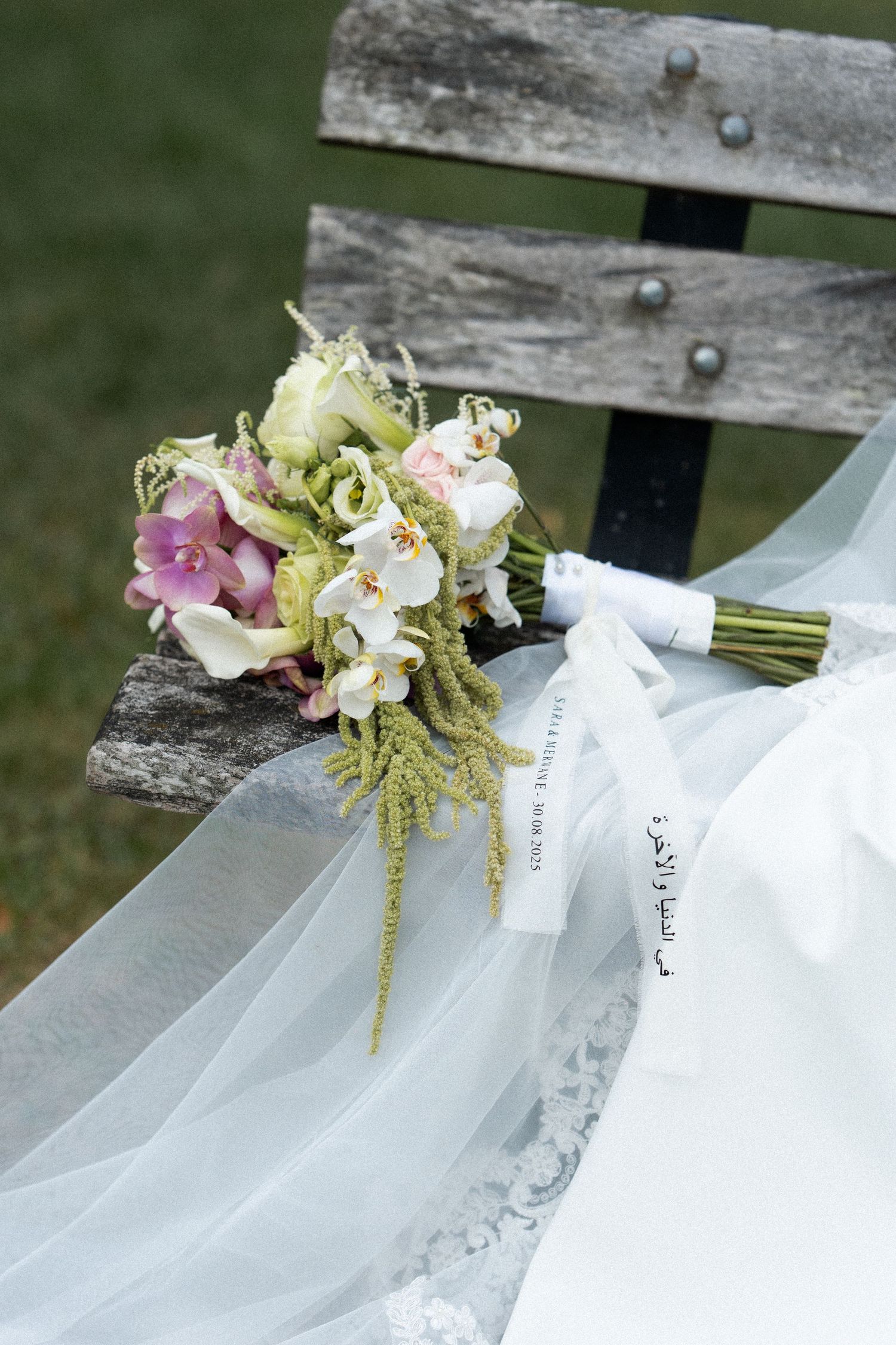 White wedding veil draped over wooden bench with delicate floral bouquet featuring orchids and greenery.