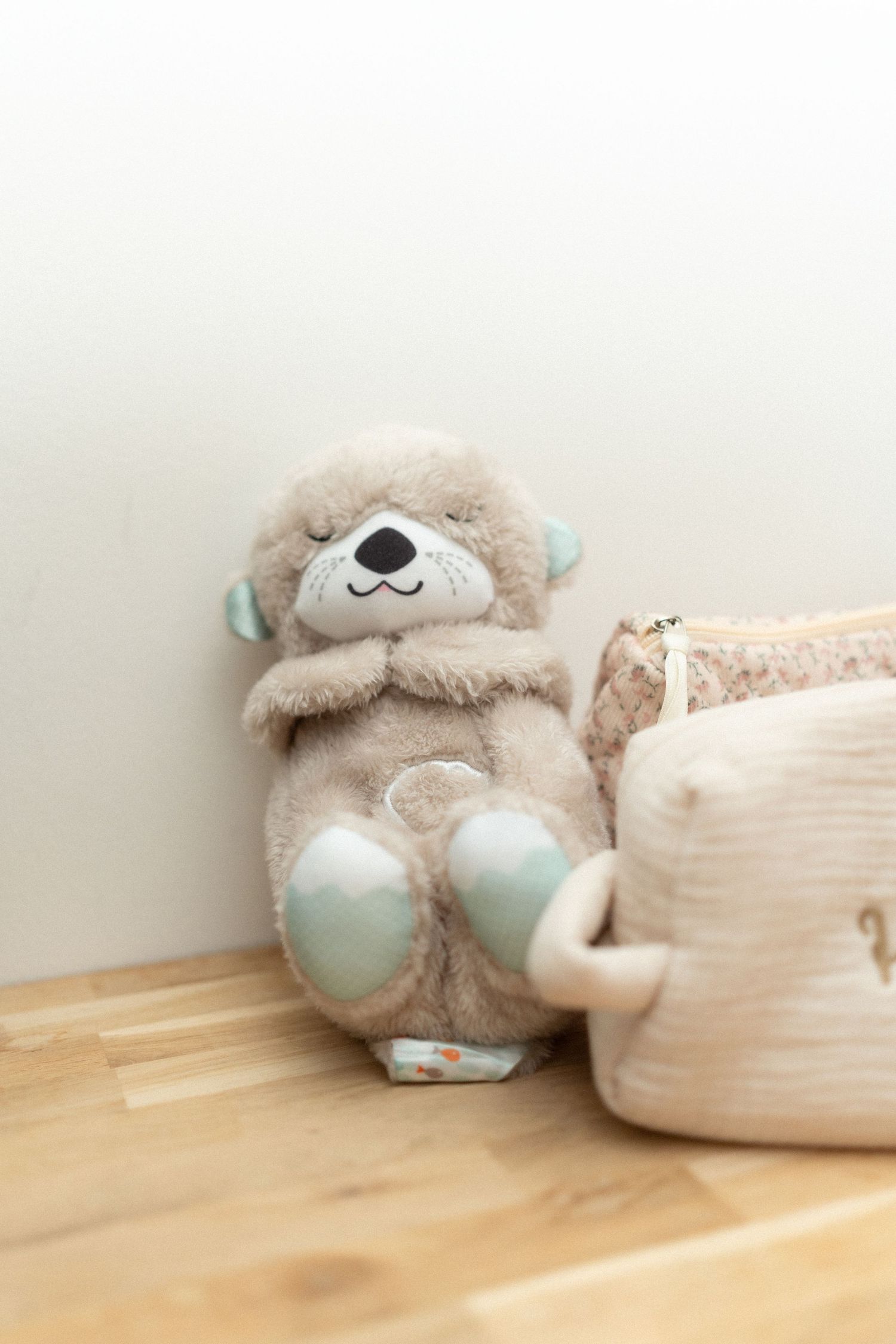 A cute plush teddy bear with mint green accents sits against a white pillow on a wooden floor.
