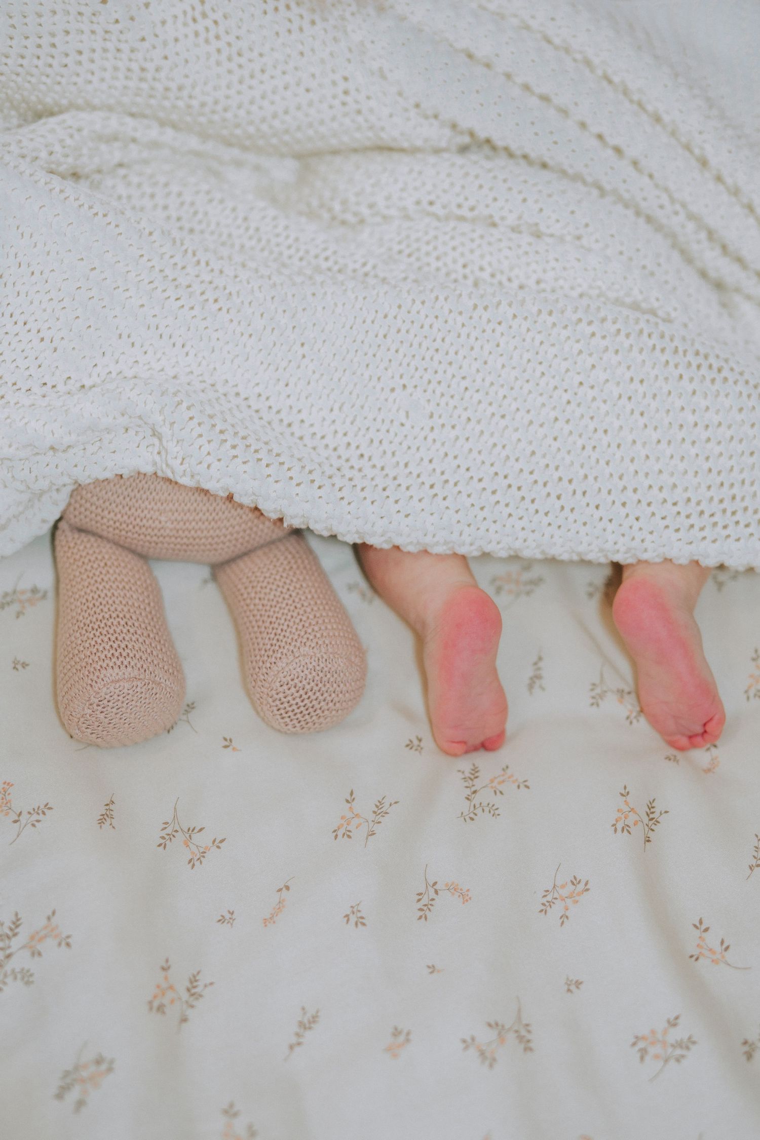 Contrasting baby feet in socks and bare feet peeking out from under a white knitted blanket.