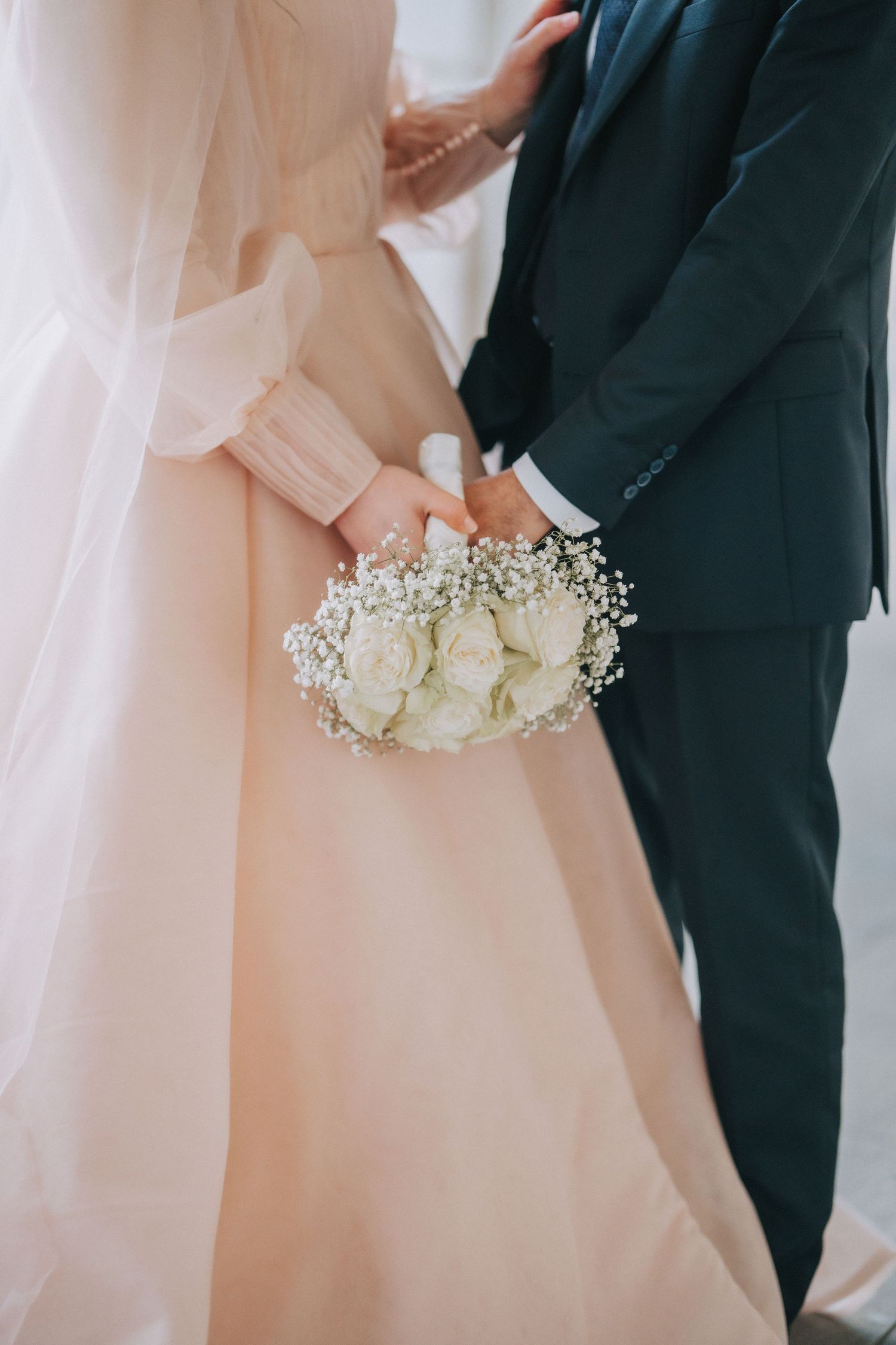 A beautiful bridal bouquet of white roses and baby's breath held against a blush pink wedding dress and dark suit.
